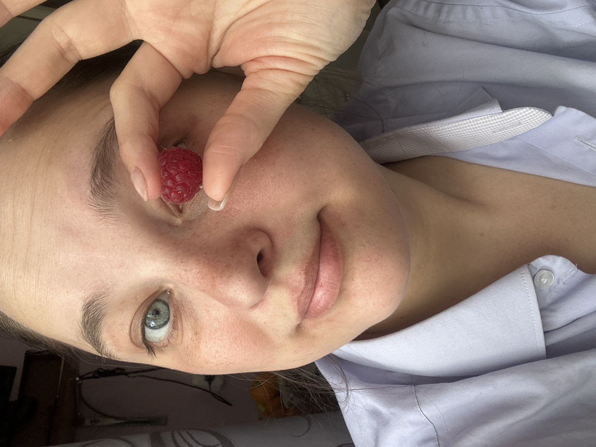 A close-up, eye-level shot shows a young person with light skin and freckles holding a single raspberry in front of their left eye, obscuring it. Their right eye is visible and is a light blue-green color. They are wearing a light purple collared shirt. The background is out of focus and appears to be an indoor setting with a shelf unit and what looks like fruit in a bowl. The image was taken in Yaroslavka, Russia.