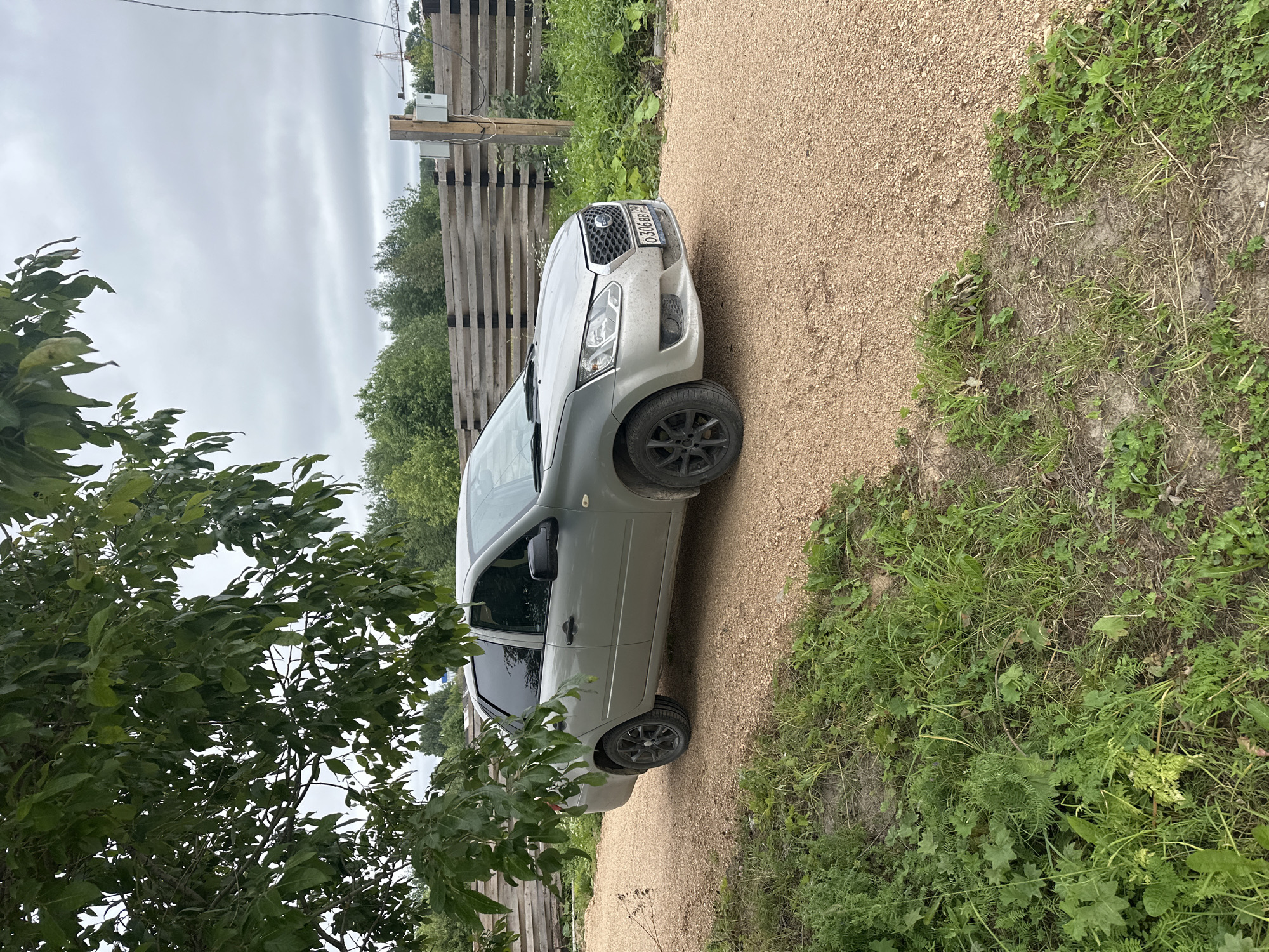 A silver sedan is parked on a gravel road in Plesetsk, Russia. The car is facing to the right, with its front slightly angled towards the viewer. The vehicle has tinted windows and black rims. To the left of the car, a large tree branch with green leaves hangs over the scene. In the background, there is a wooden fence and a dense cluster of trees under a cloudy sky. A construction crane is visible in the far distance. The ground is covered in gravel, with patches of green grass and weeds growing along the edges of the road and near the fence. The overall setting appears to be rural or semi-rural.