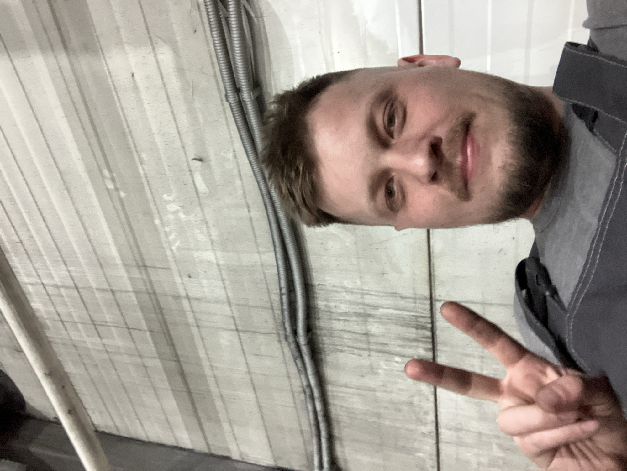 A selfie of a man with short brown hair, a beard, and wearing a gray work uniform. He is holding up a peace sign. He appears to be indoors with corrugated metal walls and some conduit in the background. There is no information to suggest that this picture was taken in Noviy, Russia.