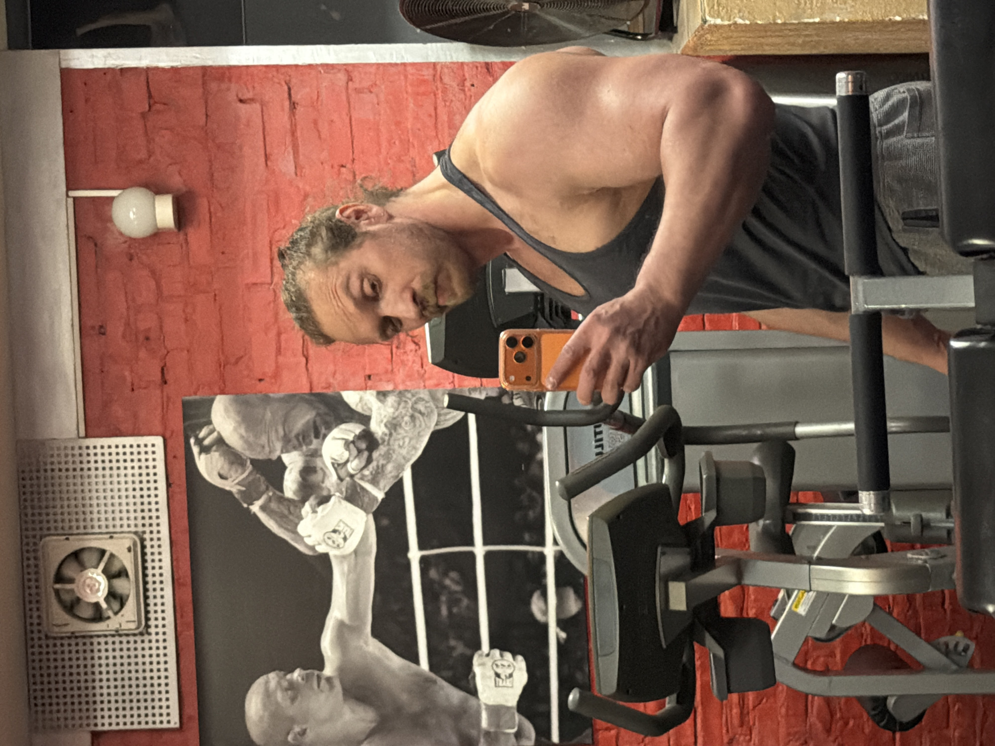 A man is in a gym, taking a selfie with an orange mobile phone. He has long hair tied back and is wearing a gray tank top. He is standing in front of a weight training machine. Behind him, there is a poster depicting two boxers in a black and white image. The walls of the gym are painted red and there is a small fan vent on the wall to the left of the poster. A light fixture is visible above the vent. The presence of the Russian writing "JITA" is present on the machine. The location in Saint Petersburg, Russia cannot be confirmed.