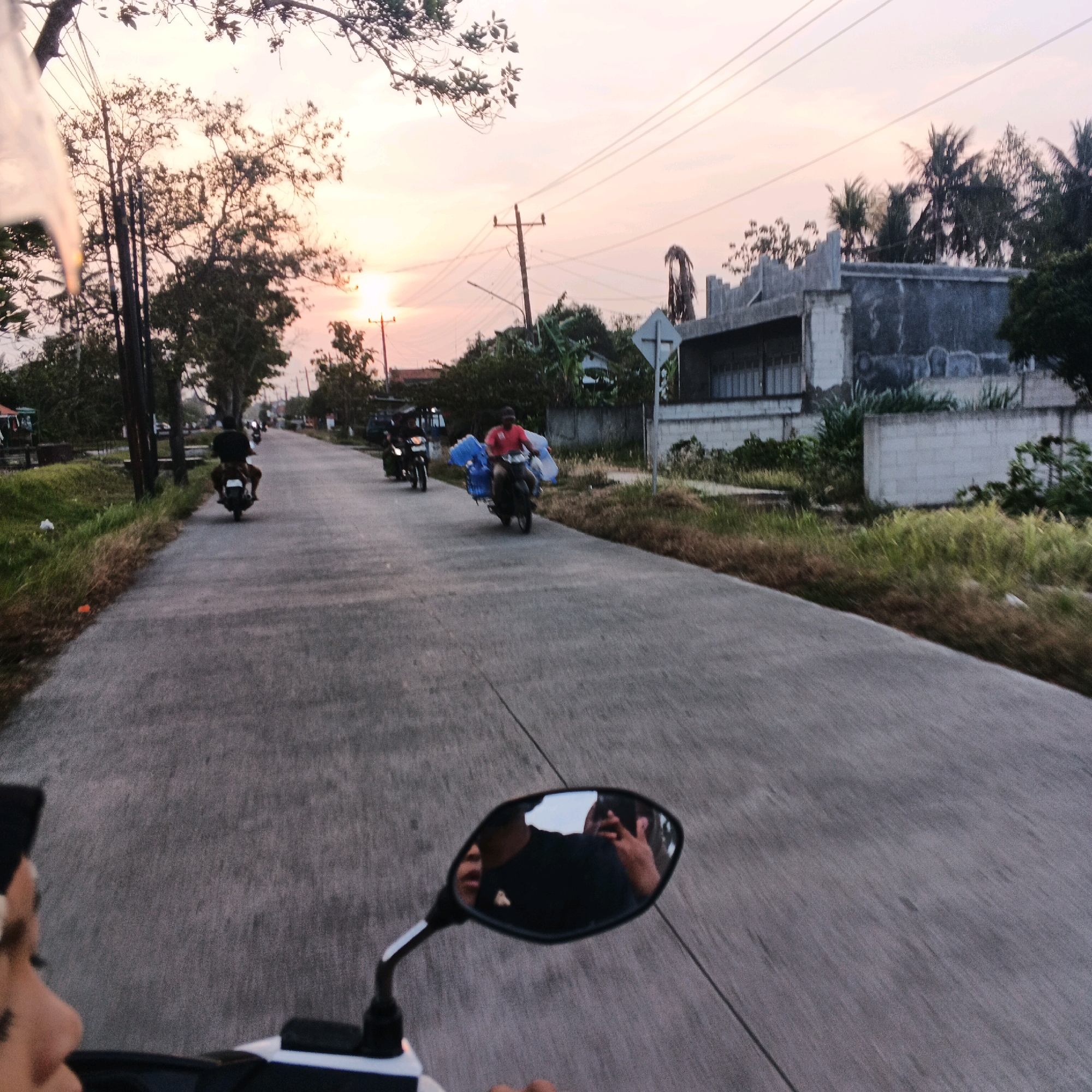 The image captures a street scene in Kalikudi, Indonesia, during what appears to be sunset or dawn. The perspective is from someone riding a motorcycle, as indicated by the motorcycle's handlebar and rearview mirror in the foreground, reflecting the rider's face and a person behind them. The road is paved and runs through a semi-rural area, with vegetation and trees lining both sides. Several other motorcycles are visible on the road, with riders engaged in various activities. One motorcycle carries large blue plastic containers, suggesting a delivery or transport of goods. Another rider is seen from behind, wearing a dark shirt. Power lines and utility poles are present, indicating infrastructure. Buildings, including a partially constructed or weathered structure with concrete walls and a tiled roof, are visible on the right side of the road. The sky displays a soft gradient of orange, pink, and pale yellow, characteristic of twilight. The overall atmosphere suggests a moment of daily life and transit in a tropical Indonesian setting.