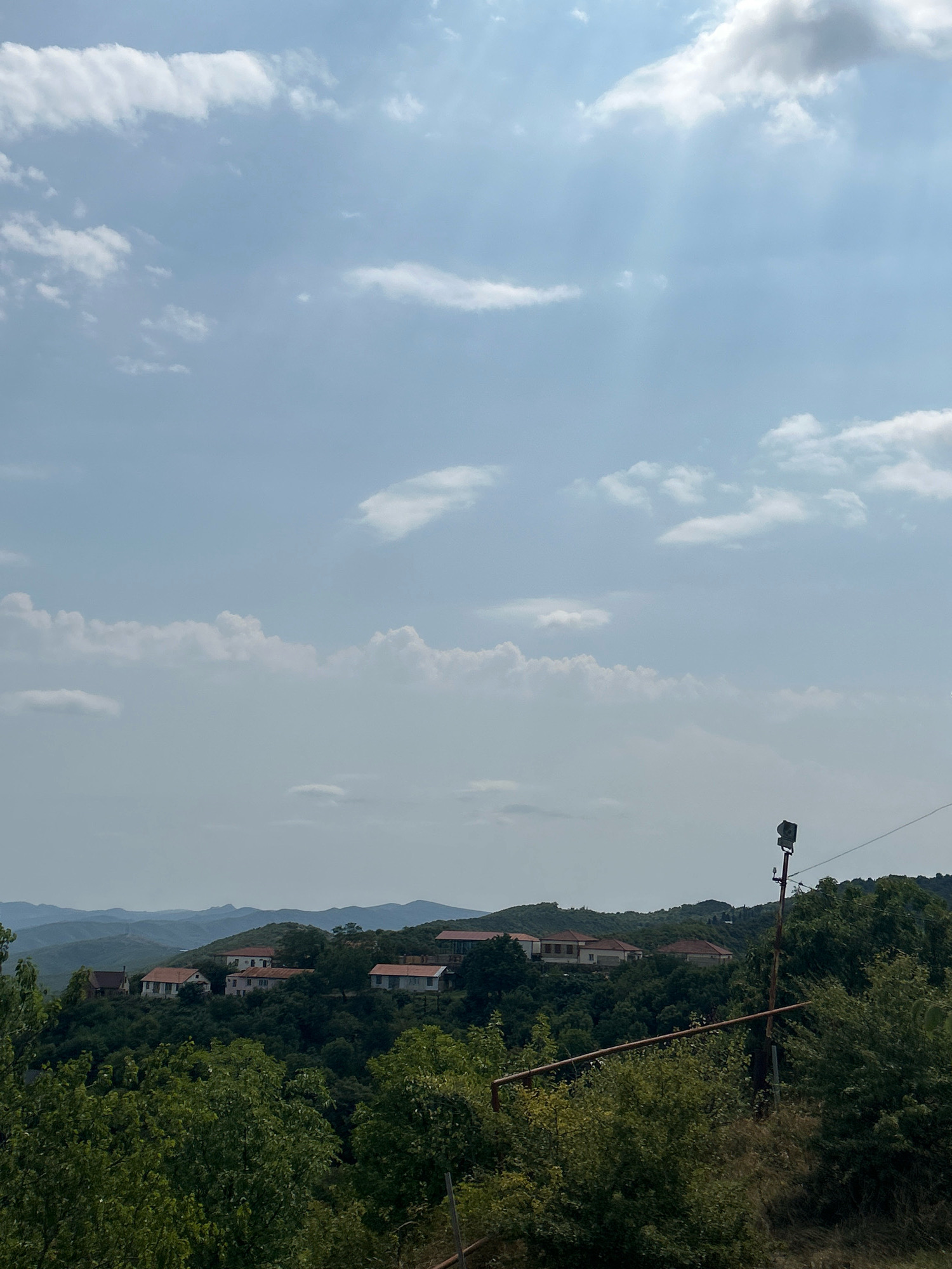 The image depicts a scenic vista of the city of Sighnaghi, Georgia. The foreground is dominated by lush green trees and foliage, suggesting a natural, possibly mountainous, environment. In the middle ground, a cluster of houses with red-tiled roofs is nestled on a hillside, indicative of a traditional settlement. The background features rolling hills and mountains, fading into a hazy horizon. The sky is partially clouded, with sunlight breaking through, casting rays across the scene. A utility pole with a light fixture is visible on the right side of the image. The overall impression is one of rural tranquility and natural beauty, characteristic of the Georgian landscape.