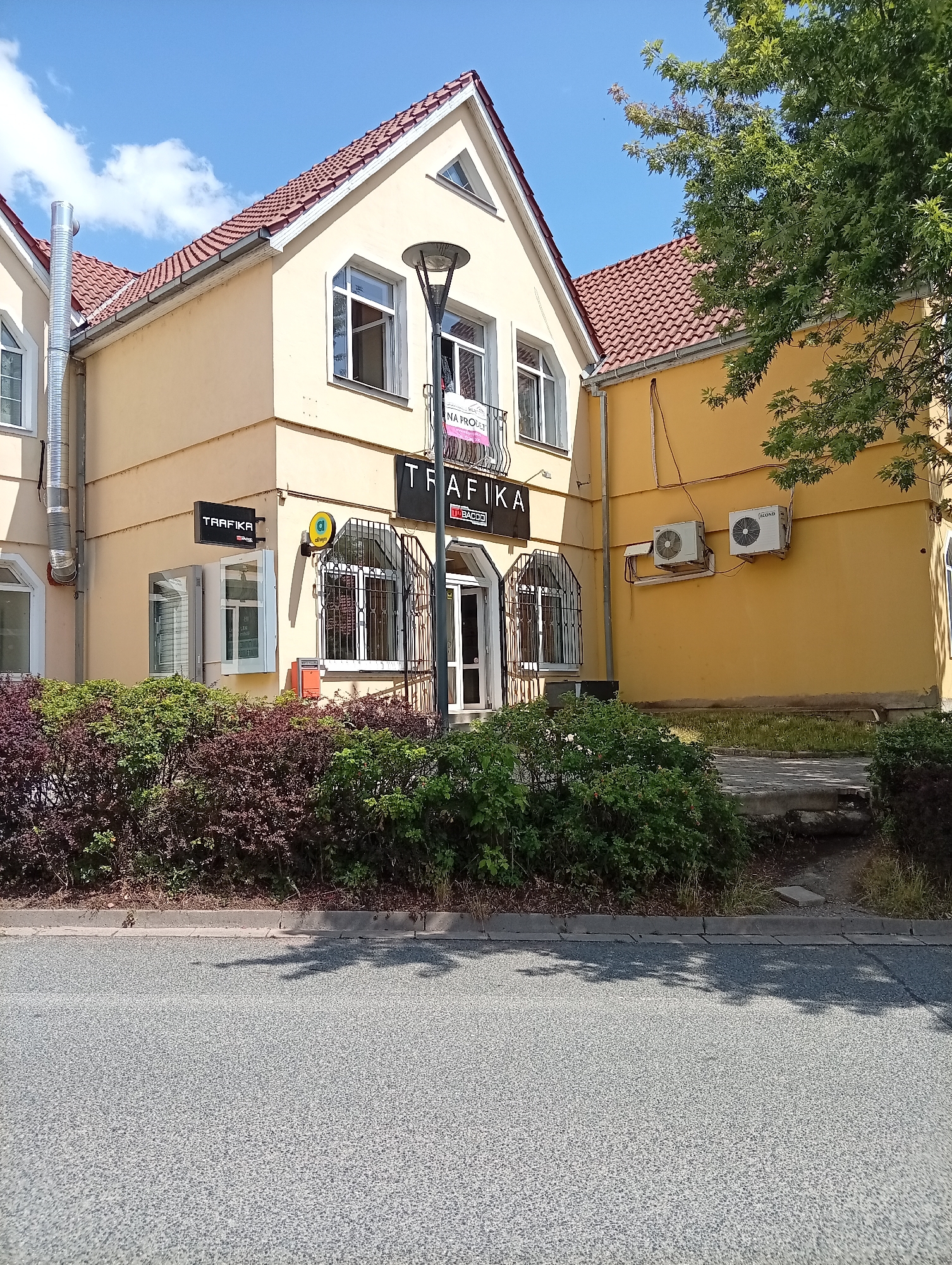 The image shows a yellow building with red tile roofing on a sunny day in the city of Tišnov, Czechia. The building has multiple windows, some with decorative metal bars. A sign on the building reads "TRAFIKA". There are two outdoor air conditioning units mounted on the yellow wall of an adjacent building. A street with a paved surface is visible in the foreground, bordered by a sidewalk and a row of green and reddish bushes. The sky above is blue with some white clouds. A mature tree with green foliage extends into the upper right corner of the frame. The scene appears to be a commercial area.