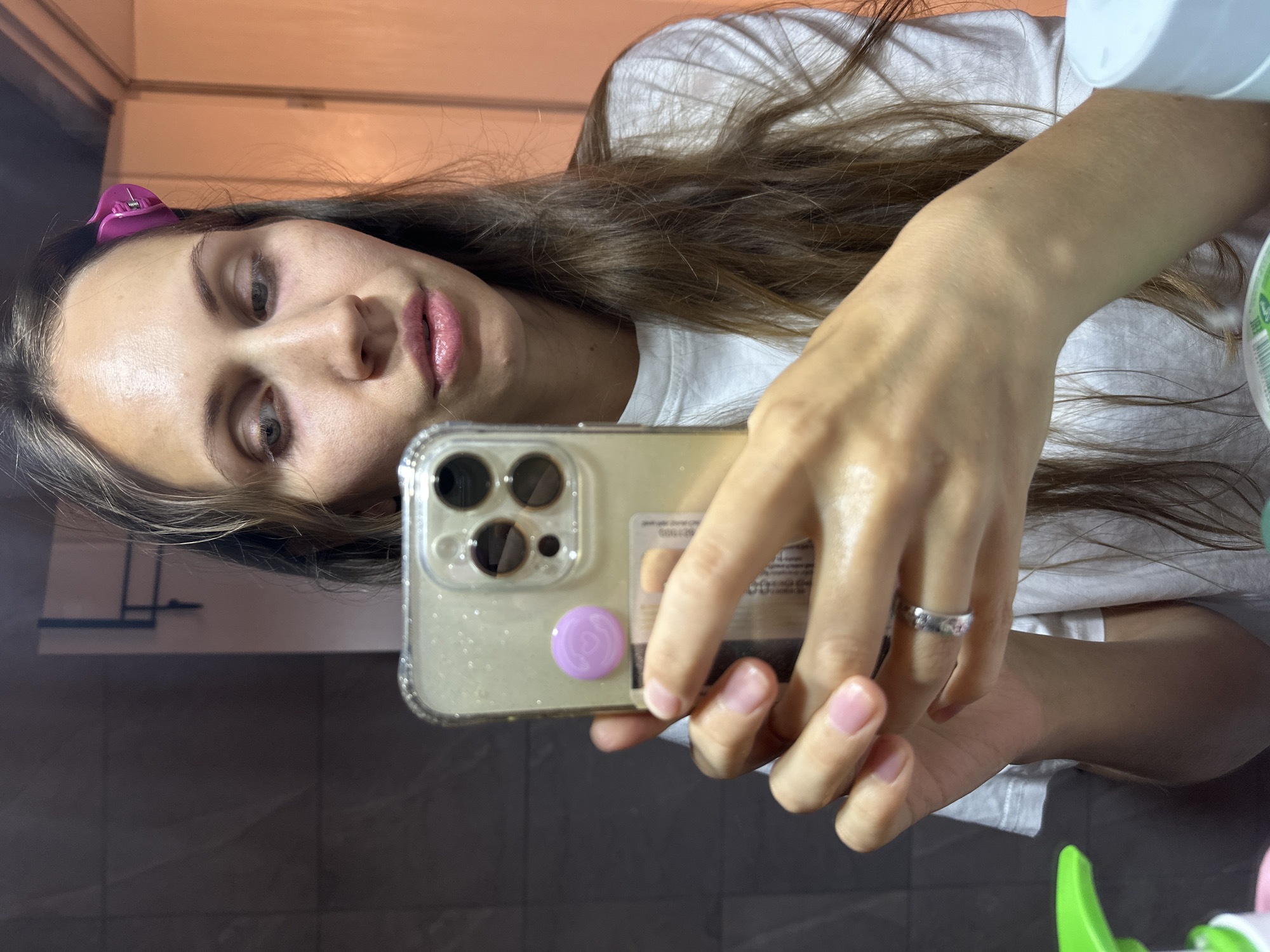 A woman is taking a selfie with a smartphone. She is wearing a white t-shirt and has brown hair with a pink clip. Her smartphone has a transparent case with a lavender-colored circular object attached. The location is indeterminate; the scene provides no information relevant to Fethiye, Turkey.