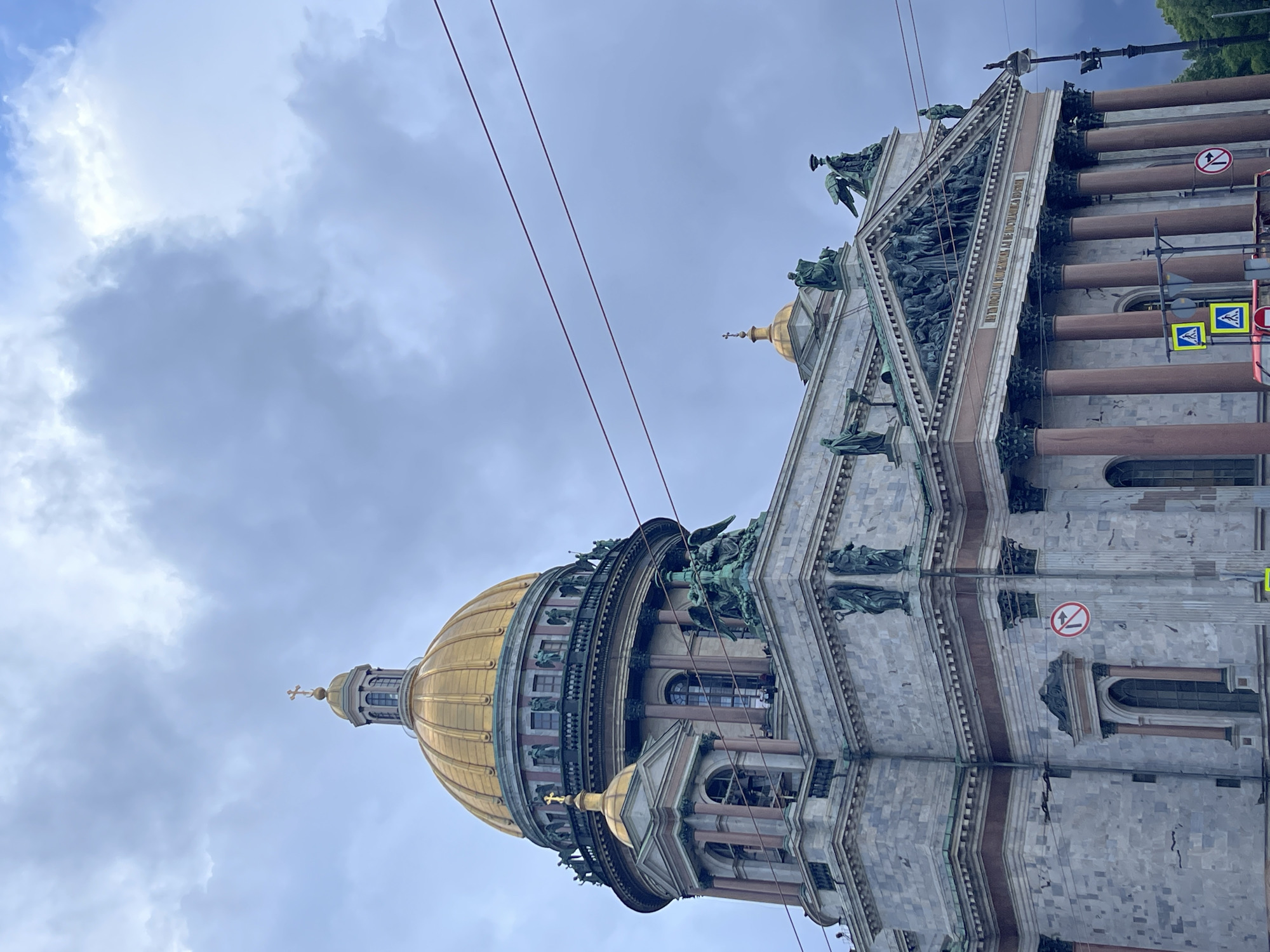 The image depicts Saint Isaac's Cathedral, a prominent landmark in Saint Petersburg, Russia. The cathedral features a massive golden dome, topped with a cross, and a smaller golden dome on an adjacent structure. The exterior is constructed of granite and adorned with statues and architectural details. Several figures, likely tourists, are visible on a balcony of the cathedral. The sky above is overcast with dramatic clouds. Power lines and traffic signs are also present in the foreground, indicating an urban setting.