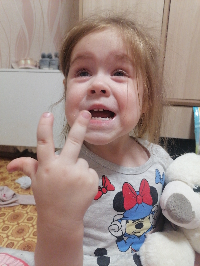 An indoor photograph depicts a young child, appearing to be a toddler, from the chest up. The child has light brown hair and an open-mouthed expression with eyes directed upwards, displaying both upper and lower teeth. The facial features are contorted, suggesting emotional distress. The child wears a light grey long-sleeved shirt featuring a graphic of Minnie Mouse in a blue hat and red bow, accompanied by smaller red and blue bow designs. The child's left hand is raised in the foreground, with the index finger extended vertically and the thumb and other fingers curled inward. Small dark spots are visible on the dorsal aspect of the child's left hand. To the right, a portion of a fluffy white stuffed animal with dark eyes and a grey snout is visible.
The background consists of a white rectangular furniture piece, possibly a low cabinet or dresser, situated on the left, with two pairs of small footwear (one light-colored, one grey) on its top surface. Behind this furniture, a light-toned wallpaper with a repeating, undulating abstract pattern is present. The floor visible beneath the child and to the left is covered in patterned linoleum or vinyl flooring in shades of brown and yellow, with a crumpled piece of light-colored fabric resting on it. A vertical section of light-colored wood paneling or a door frame is visible on the far right, alongside a blue and white striped object. The specific location within the city of Chaikovsky, Russia, cannot be determined from the visual information provided.
