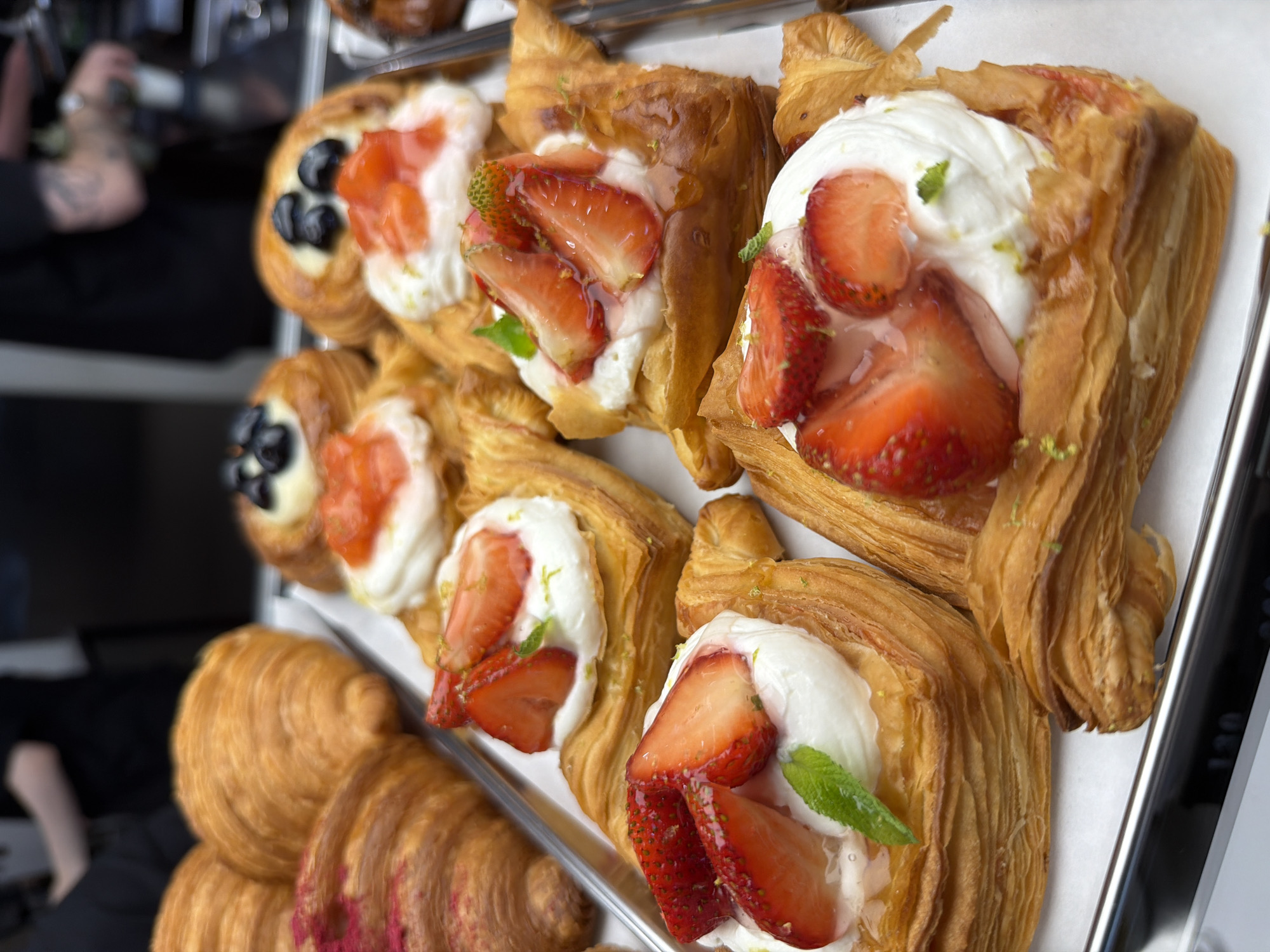 A close-up, eye-level shot captures a tray of fresh pastries, likely at a bakery or cafe. The pastries are arranged on a white surface, with a metal tray visible at the bottom edge of the frame. In the background, out of focus, a person in a dark shirt is visible, suggesting a commercial setting. The pastries themselves are the main focus. There are several flaky croissants, some topped with a pink dusting. Adjacent to the croissants are danishes topped with dollops of white cream, fresh strawberries, and what appears to be a light green zest, possibly lime. One danish also features blueberries. The lighting is bright, highlighting the golden-brown crust of the pastries and the vibrant red of the strawberries. This scene is not identifiable as being in Izhevsk, Russia.