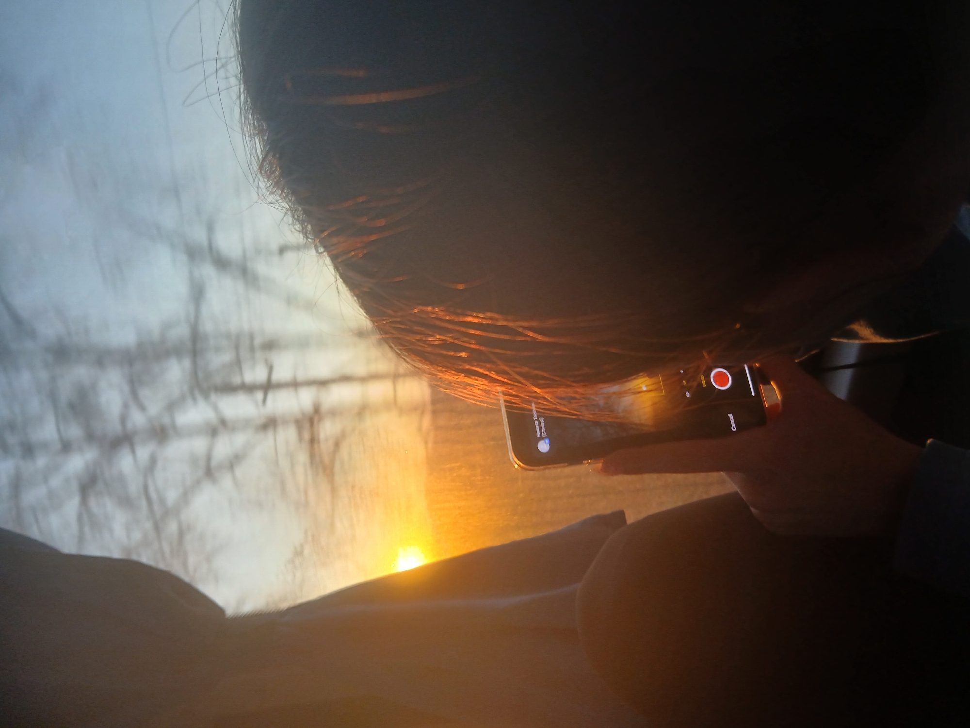 A close-up, rotated image shows a person recording video with a smartphone. The person's dark hair is visible in the foreground, illuminated by the sunlight. A hand holds the smartphone, and the phone screen displays recording controls. In the background, a bright sun shines on a blurred landscape, possibly taken from inside a vehicle. The sky appears hazy or foggy, with silhouettes of bare trees visible. There is no information in the image to confirm the location as Osa, Russia.