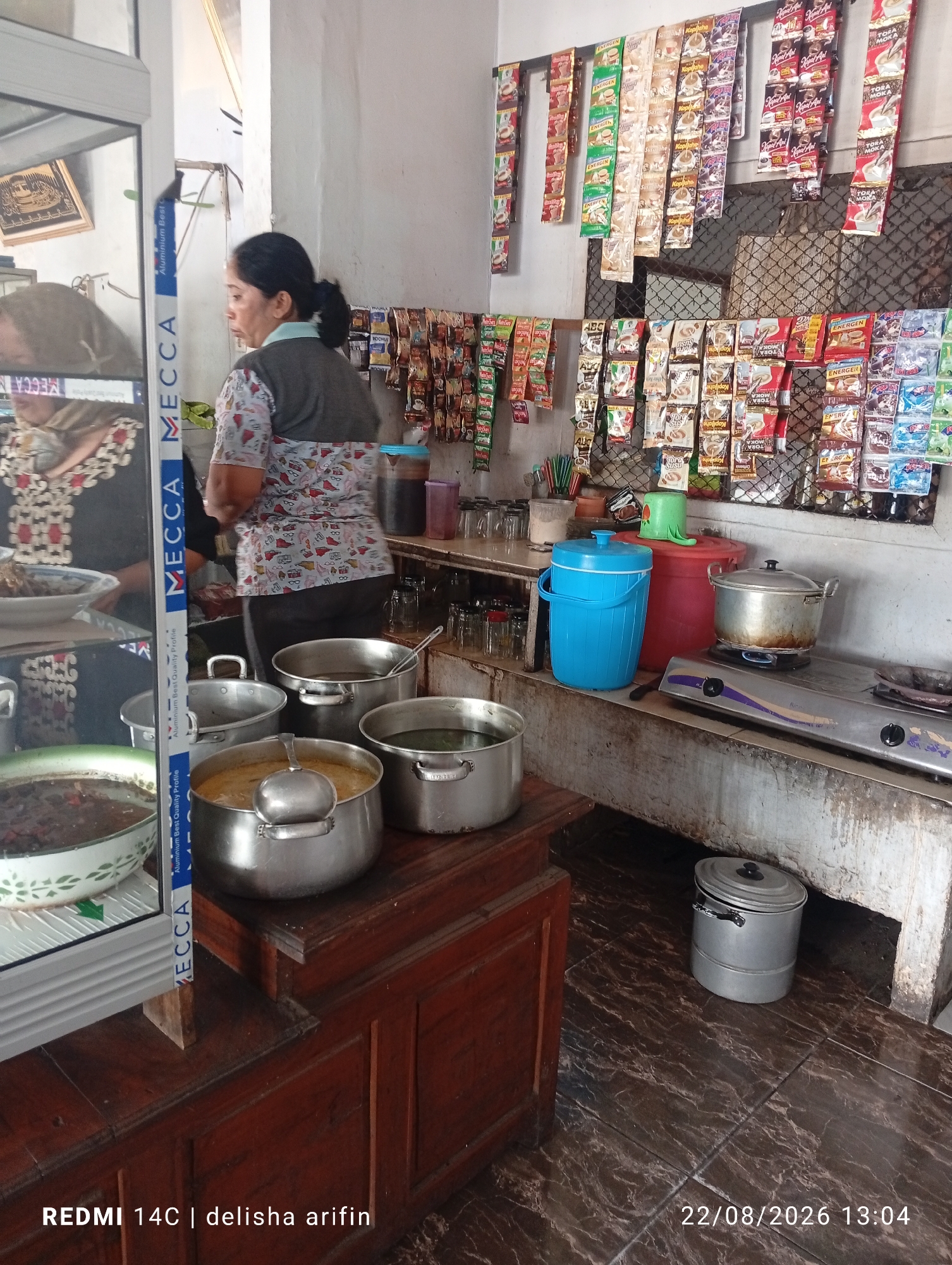 In a dimly lit eatery, likely a small warung in Gambir Kuning, Indonesia, a middle-aged woman with dark hair pulled back stands behind a counter, tending to food preparation. She wears a grey vest over a patterned shirt and dark pants. In front of her, a stainless steel stove holds a pot of simmering liquid, and to its right, a blue cooler and a red container are visible. Several large pots, some containing food and others empty, are arranged on a wooden surface. A display case on the left shows a bowl of food. Above the counter, shelves are stocked with numerous individually packaged coffee and beverage sachets, hanging in rows. The floor is tiled with a dark, patterned design. A metal bin sits on the floor to the right. A camera timestamp in the lower right corner reads "22/08/2026 13:04".