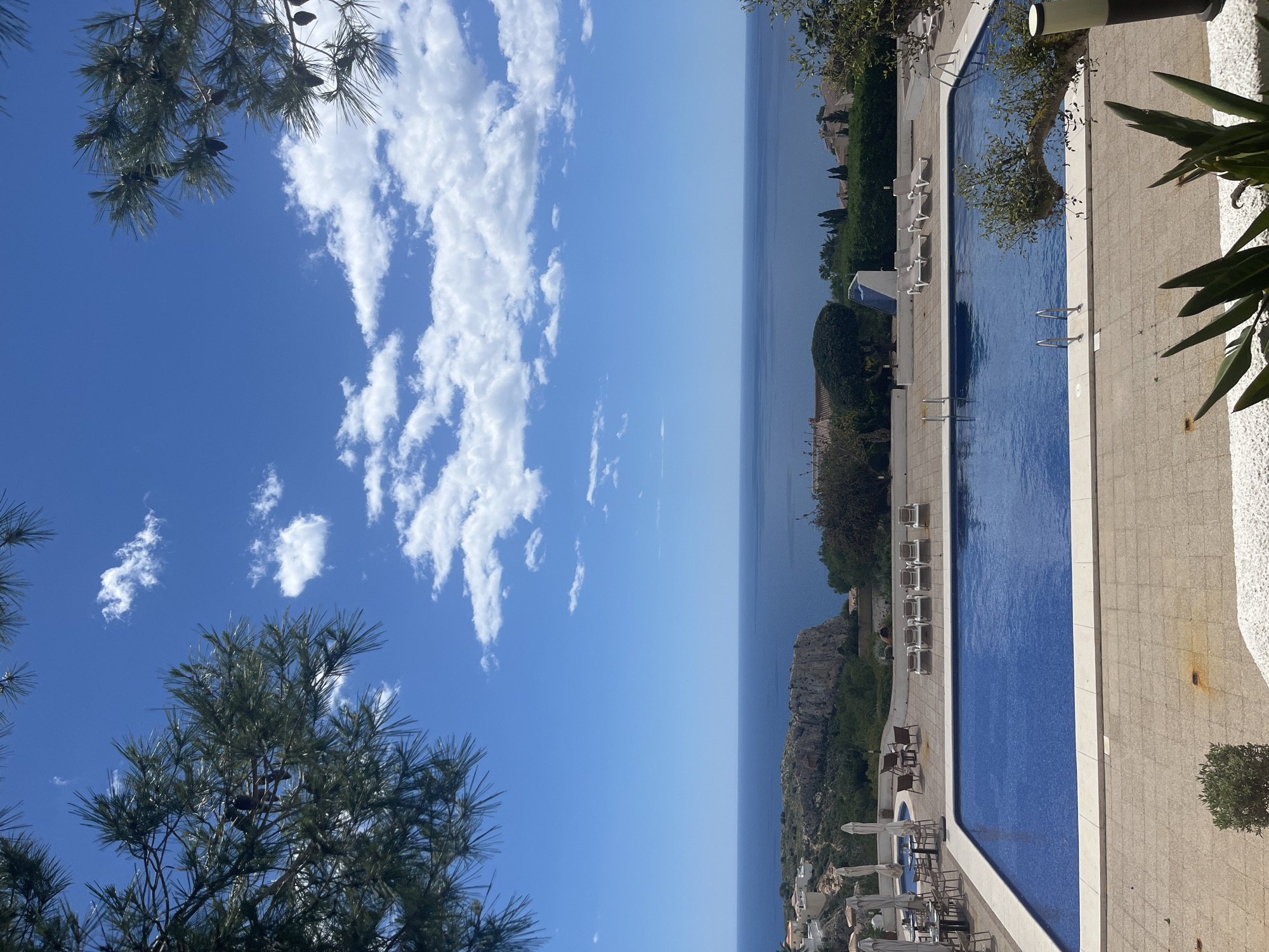 The image captures a view from a high vantage point in El Poble Nou de Benitatxell / Benitachell, Spain. The scene features a clear blue sky with scattered white clouds on the left. Below the sky is a horizontal band of blue sea meeting the horizon.
To the right, a rectangular swimming pool filled with blue water dominates the foreground. The pool is surrounded by a light-colored tiled patio area with sun loungers positioned along the side, some with umbrellas. The coastline and hillside are visible in the background, suggesting a coastal location. Green vegetation is interspersed throughout the scene, including trees along the edge of the frame. The photo seems to have been taken during daylight.