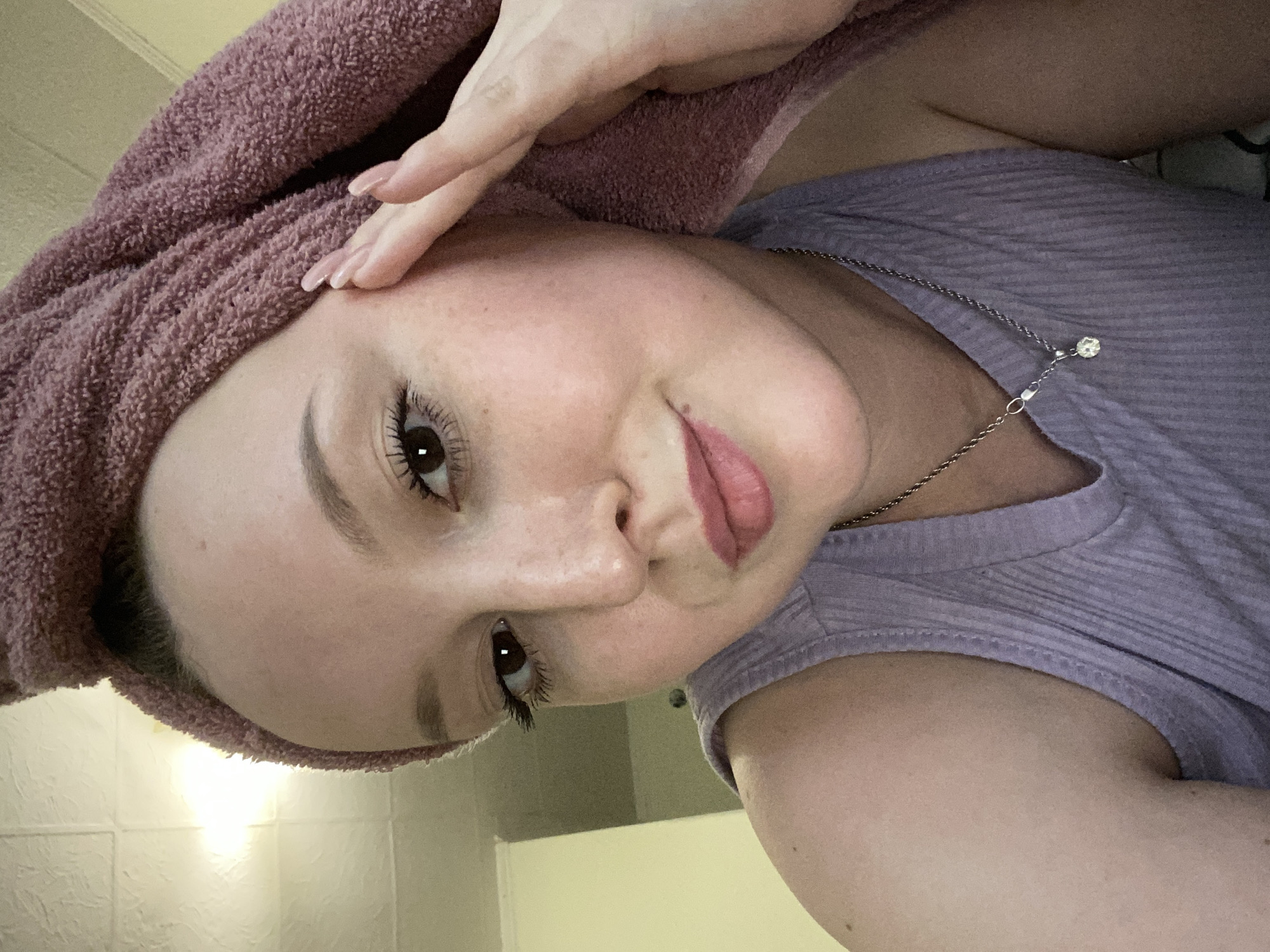 A person with a towel wrapped around their head is looking at the camera. They are wearing a lilac tank top and a silver necklace with a pendant. Their left hand is raised to their forehead. The background is a pale yellow and white wall. There is no indication of the location being Chaikovsky, Russia.