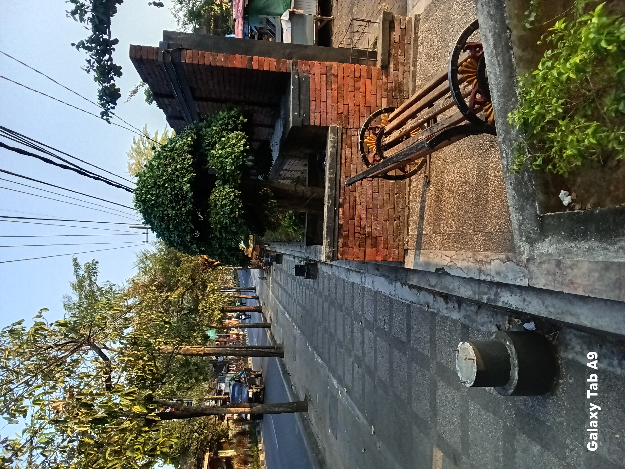 The image depicts a street scene in Kelutan, Indonesia. A sidewalk with a patterned stone surface runs parallel to a paved road. Trees with green foliage line the sidewalk, casting shadows. A black pickup truck and a motorcycle are visible on the road. On the right side of the sidewalk, there is a brick structure with a decorative element resembling a wooden cart or bench with spoked wheels. A neatly trimmed green bush is planted in front of this structure. Overhead, power lines stretch across the clear blue sky. The scene appears to be captured during daylight, possibly late afternoon, given the warm lighting.