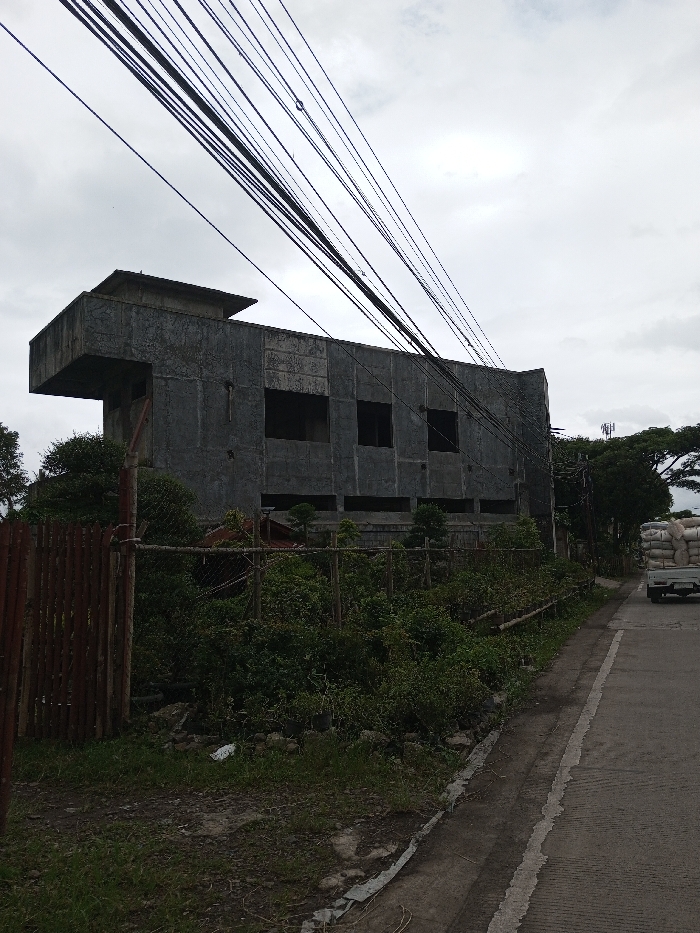 The image displays an outdoor scene in Melakudikadu, India, under an overcast sky. A multi-story concrete structure, appearing unfinished with exposed grey concrete walls, dominates the left-center. The building features multiple rectangular window openings without glass and a distinctive cantilevered section on its left side. In the foreground, a fence composed of vertical wooden planks and wire mesh separates a dense area of green vegetation and dirt from a paved asphalt road. Numerous black utility lines crisscross diagonally across the upper portion of the frame. On the far right side of the road, a light-colored pickup truck is partially visible, loaded with several large white sacks, and appears to be moving away from the viewer. Beyond the road and vehicle, trees and a utility tower are present in the background. No individuals are visible.