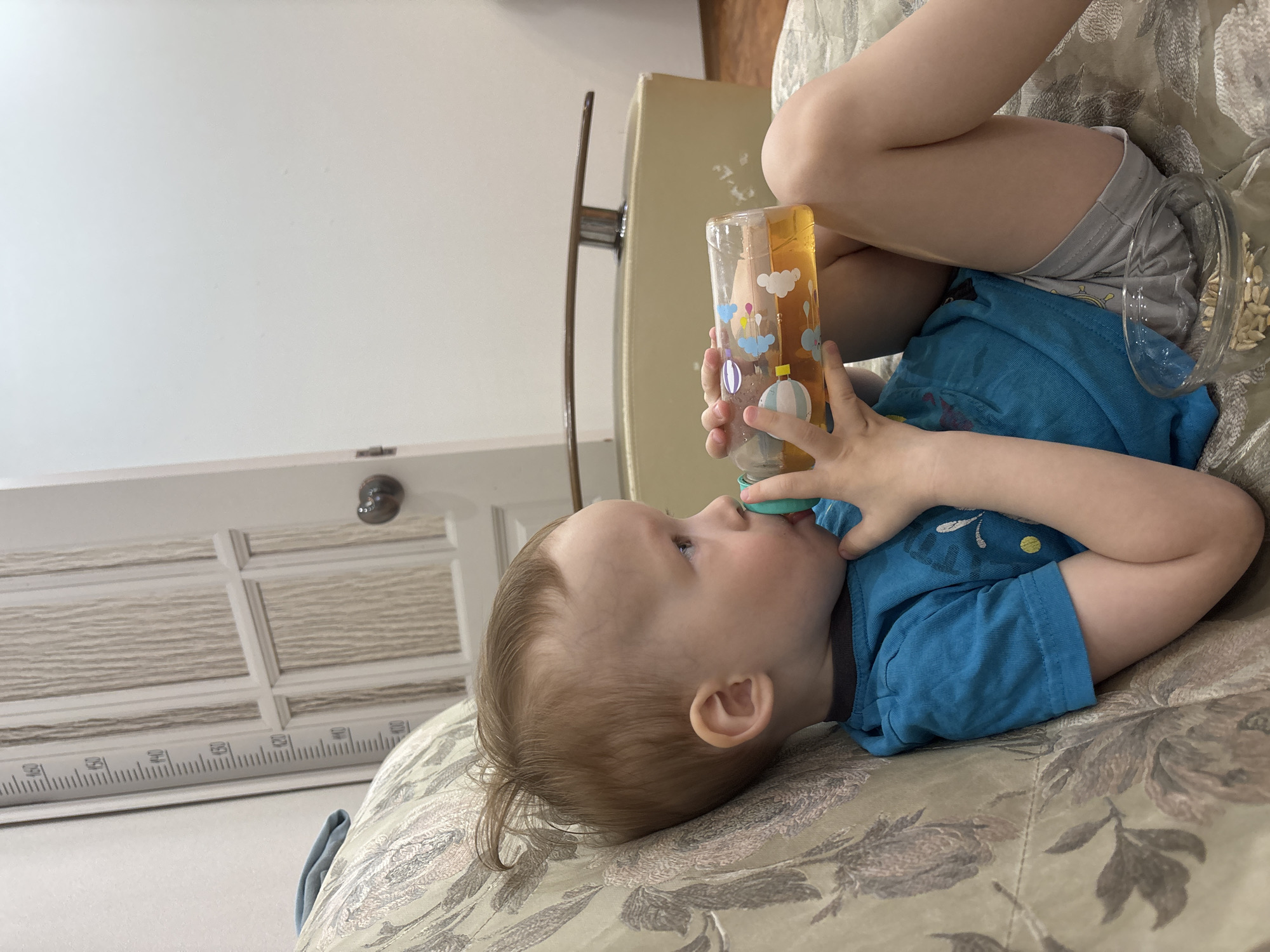 A child is lying on a patterned sofa while drinking from a plastic bottle with hot air balloon designs. The child is wearing a blue shirt and gray shorts. The sofa is upholstered with a beige and brown floral pattern. In the background, a white door with decorative panels and a silver doorknob is visible. A height chart is attached to the doorframe. A light-colored piece of furniture, possibly a cabinet, with a metal handle is positioned behind the child. The location is Ферма, Russia.
