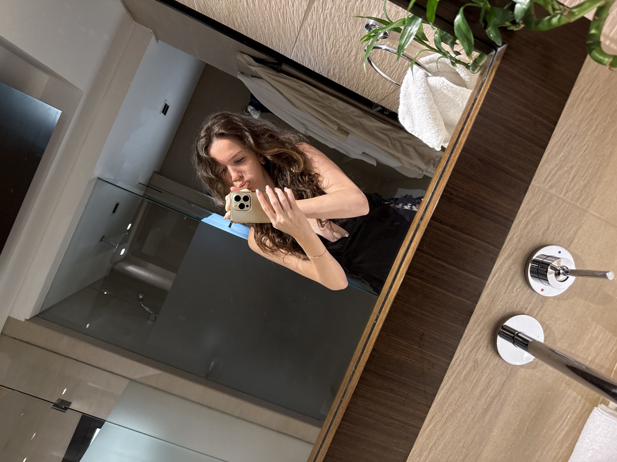 A young adult female, with long, wavy brown hair, is taking a mirror selfie in a modern, well-lit bathroom, consistent with luxury accommodation in Dubai, United Arab Emirates. She is wearing a black, low-cut top and holds a gold-colored smartphone with a triple camera array in her right hand, positioned to capture her reflection. Her left hand is also visible, with light-colored manicured nails. She is making a puckered lip expression towards the camera. A delicate gold bracelet is visible on her right wrist.
The bathroom features a large mirror with a dark wood frame at the base, reflecting a light beige tiled wall directly behind the woman. To the left, a clear glass shower partition with a frosted section is visible, revealing a dark interior beyond, possibly the shower area with a chrome shower head and other fixtures. The ceiling is white with recessed lighting. In the foreground, below the mirror, is a light-toned wooden counter with two modern chrome sink faucets; one is a single lever, and the other has a circular handle with red and blue indicators. A white, folded hand towel rests on a chrome towel ring to the right of the faucets. Further right, a small potted green plant, identified as Lucky Bamboo, is placed on the counter. The floor is covered in light-colored tiles. A beige robe or towel hangs on hooks reflected on the far right wall.