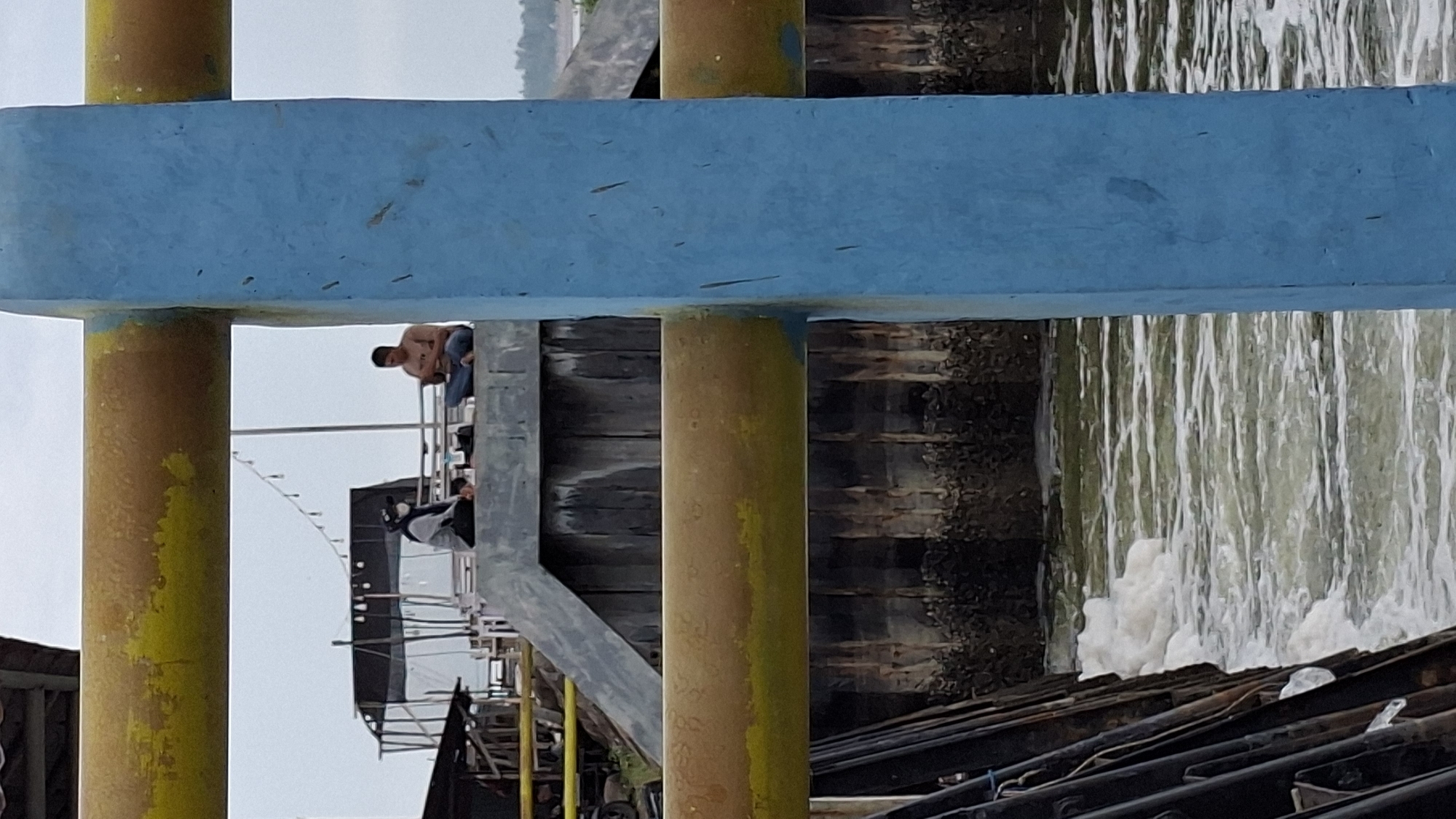 The image shows two people sitting on a concrete structure next to a body of water. One person, wearing a headscarf and mask, is seated with their legs crossed. The other person is also seated with their legs crossed and appears to be looking at something in their hands. In the background, there are some structures that suggest a waterfront area, possibly a pier or a jetty. The water has white foam on its surface, indicating movement. The scene is framed by a yellow and a blue railing. The location is identified as Lhokseumawe, Indonesia, suggesting it is a public space by the sea or a river.