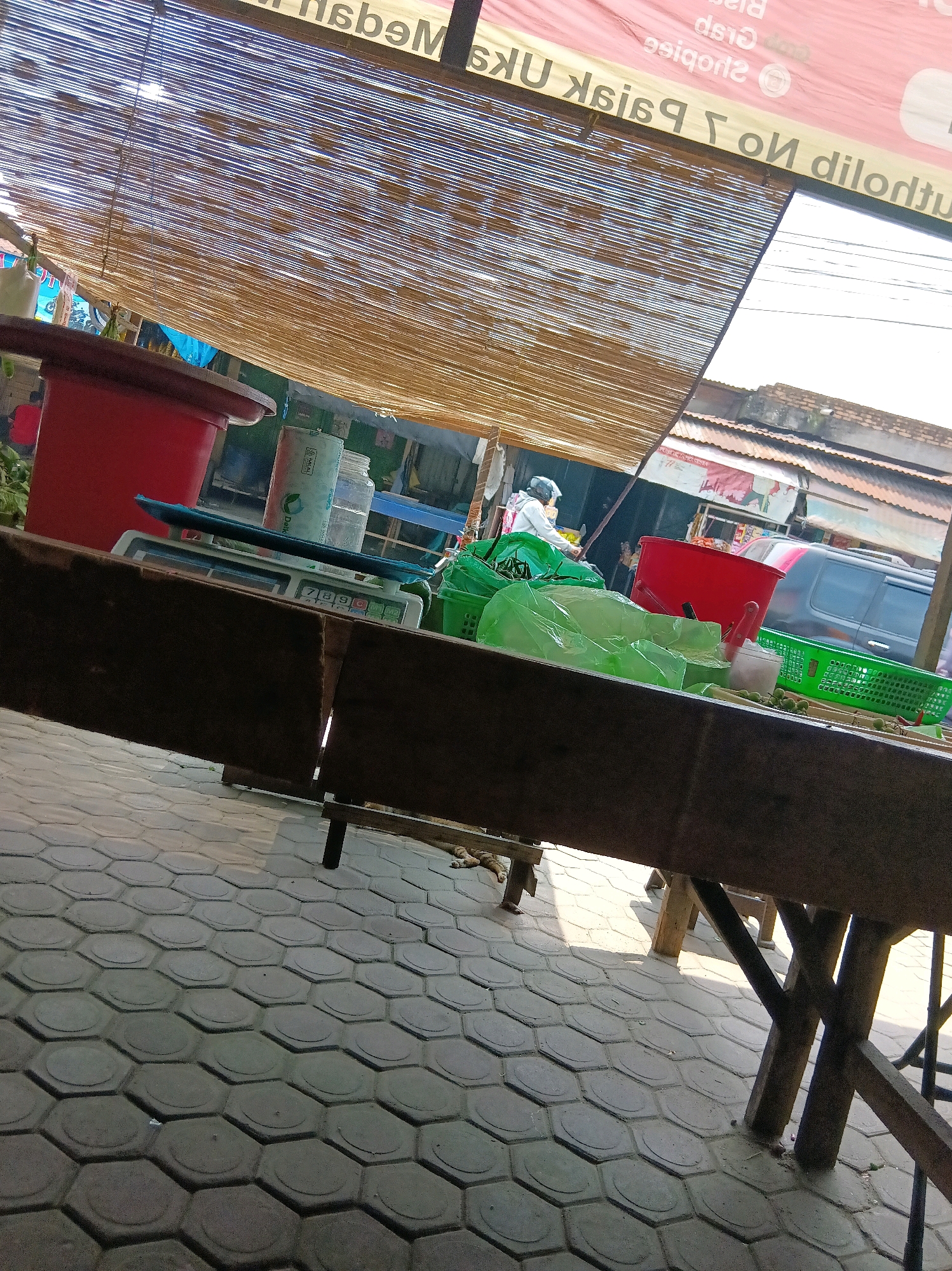 This is a street-level shot taken in the City of Medan, Indonesia. The scene appears to be a market stall under a woven bamboo canopy. In the foreground, a rustic wooden table occupies the lower portion of the frame. Beyond the table, a red plastic tub, a digital scale, and several green plastic bags are visible. A person wearing a white shirt and helmet is seen riding a motorcycle in the background. The ground is paved with hexagonal grey tiles. The overall impression is one of a bustling outdoor market.