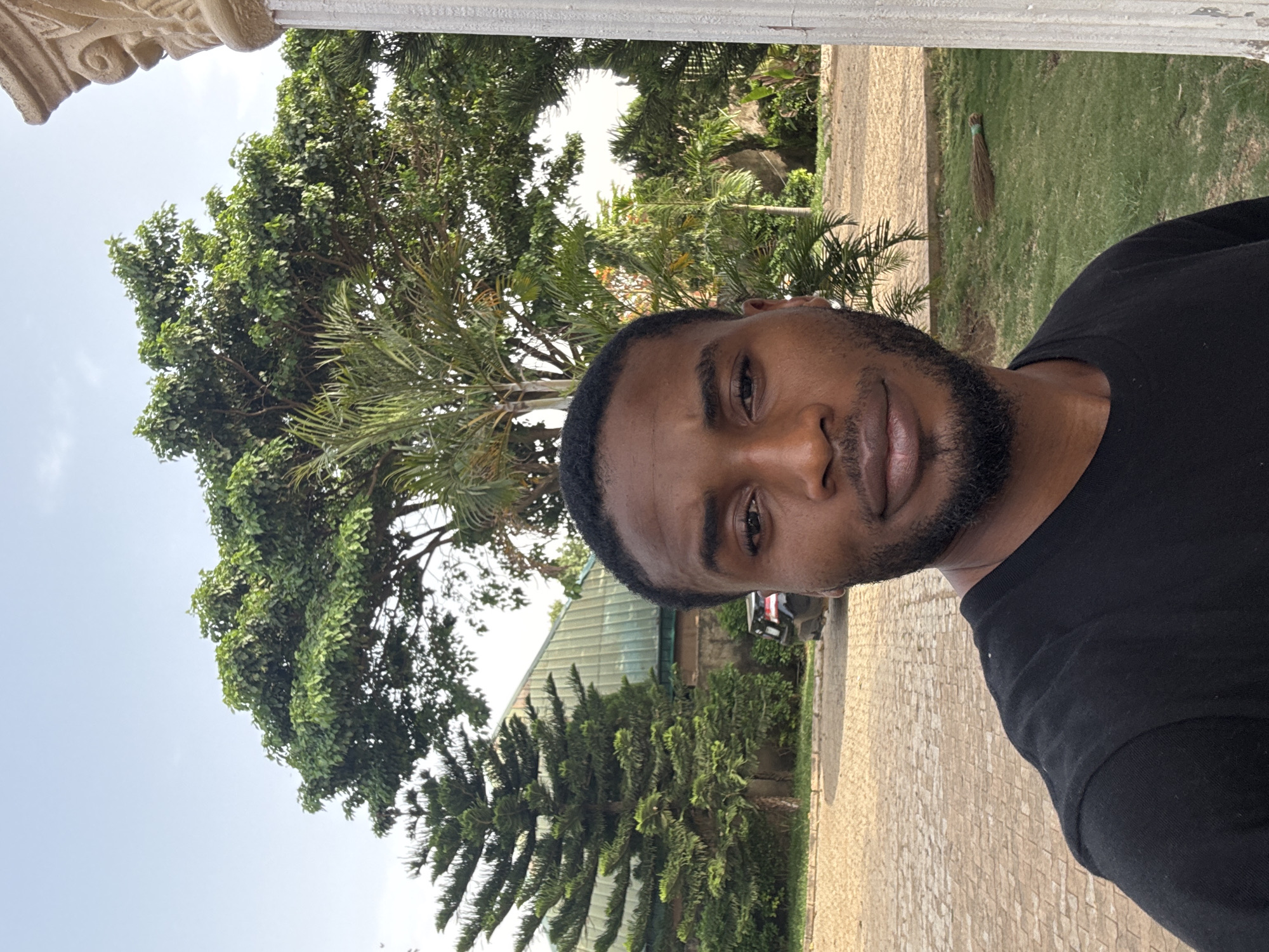 A medium close-up shot features a dark-skinned man wearing a black shirt, with his head and upper torso visible. He has short dark hair and a beard. The background shows a mix of trees and vegetation. A building with a visible roof is also in the frame. A paved area is seen on the right side of the image. Based on available information, the location is Jos, Nigeria. A broom can be seen standing on the grass.