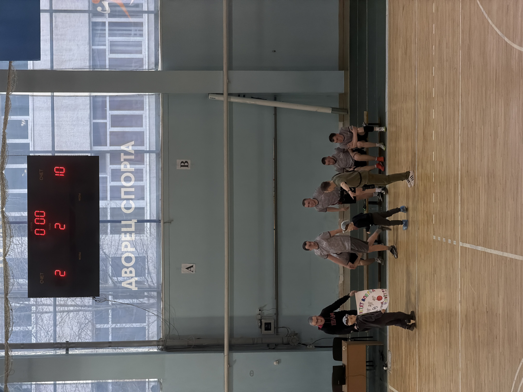 Here's a forensic description of the image:
**Scene:** An indoor gymnasium, potentially in Konratovo, Russia, based on the OCR of Cyrillic text.
**Environment:** The gym has a wooden floor with lines indicating a sports court (likely basketball or volleyball). Large windows provide daylight. The walls are light-colored, with column markers labeled "A" and "B".
**Objects:**
* An electronic scoreboard displays "2" for the home score, "10" for the away score, "2" for period, and "0:00" time.
* A banner is held by a person.
* A row of chairs are placed along one side of the court.
**People:**
* A group of people are standing or performing exercises on the court. They appear to be young men, some wearing athletic clothing.
* One person is holding a banner, wearing a hat, and a "TEAM" shirt.
* Other men are seated on benches, facing the court.
**Actions/Interactions:**
* Some of the men are engaged in what appears to be a stretching or warm-up routine.
* The person holding the banner is facing the camera.
**Overall Impression:** The image captures a scene in a gymnasium, possibly during a break or warm-up period of a sports event.