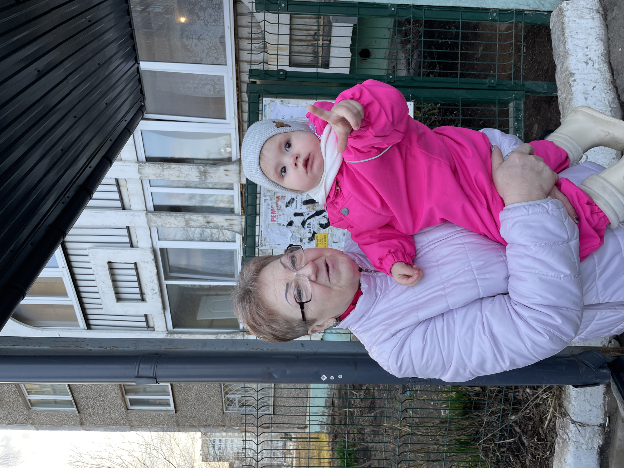 An elderly woman holds a baby in her arms while standing outdoors near a building. The woman is wearing glasses and a light purple puffy jacket. The baby is wearing a pink snowsuit and a light gray hat, pointing their finger forward. They are standing near a green metal fence. Behind them, there is a building with multiple windows and a black metal structure above. Based on visual markers, this scene is likely located in Izhevsk, Russia.