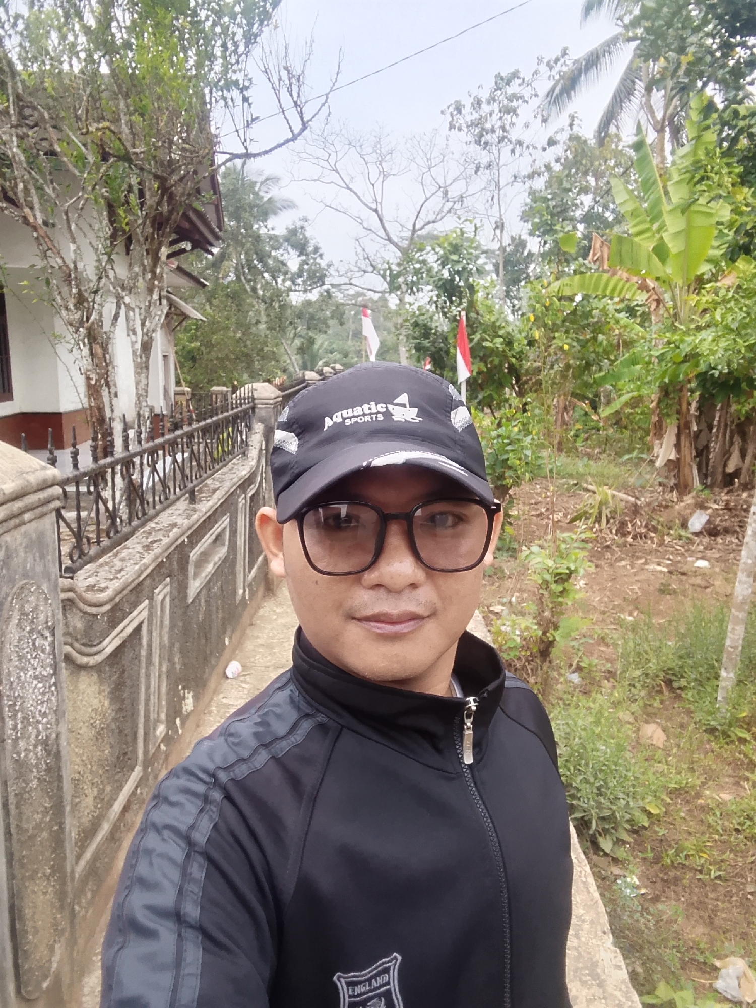 A close-up, eye-level, outdoor selfie shows a person wearing a black jacket and a black baseball cap with the text "Aquatic SPORTS" and a small logo. The person is also wearing dark-rimmed glasses. Behind the person, a concrete wall with decorative iron railings runs alongside a dirt path. Beyond the wall and path, a lush green environment with trees, foliage, and what appears to be banana plants is visible. A white and red flag, possibly the Indonesian flag, is seen in the background among the trees. A white building with a brown roof and some trees are also visible to the left. The overall impression is a rural or semi-rural setting.
