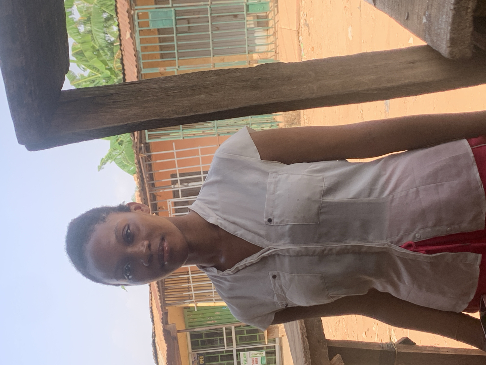 A woman stands under a wooden structure in Sabon Tasha, Nigeria. She is wearing a white, button-down shirt and a red garment. In the background are buildings with metal roofs and barred windows. The sky is visible in the upper left corner.