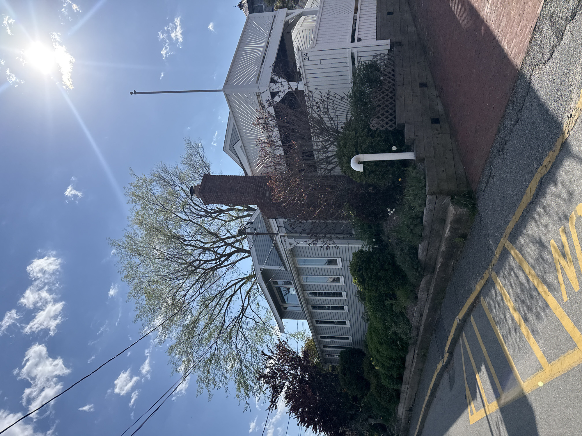The image shows a building in Provincetown, Massachusetts, United States. The building is a two-story structure with gray siding and white trim. There is a red brick chimney on the side of the building and a white pergola extending from the back of the building. The number "24" is visible on the pergola. A large tree with sparse leaves stands in front of the building. The sky is blue with some scattered white clouds, and the sun is shining brightly. A street with yellow markings is visible in the foreground. Power lines run across the sky in the lower left of the frame. The image has been rotated, and the scene appears angled to the right.