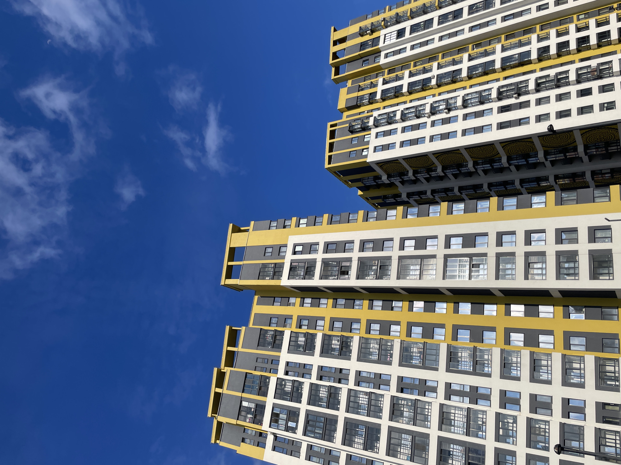 This low-angle, upward-looking photograph showcases a striking, modern multi-story building against a clear, vibrant blue sky dotted with scattered white clouds. The building, located in Izhevsk, Russia, exhibits a distinctive architectural style characterized by a bold combination of bright yellow, dark gray, and white panels.
The structure features a series of cantilevered and staggered sections, giving it a dynamic, almost sculptural appearance. Numerous windows, mostly rectangular or square, punctuate the facade, with some reflecting the bright sky. A notable decorative element is the intricate circular pattern in yellow and black visible on the underside of several prominent overhangs or balconies. Below some windows, small, dark external fixtures, possibly air conditioning units, are mounted. A black light fixture or camera can also be observed on one of the white wall sections.
In the upper-left quadrant of the blue sky, a faint crescent moon is visible, indicating that the photograph was taken during daylight hours. The overall impression is one of clean lines, geometric precision, and a modern aesthetic. There are no people or discernible activities taking place, and no visible text in the image.