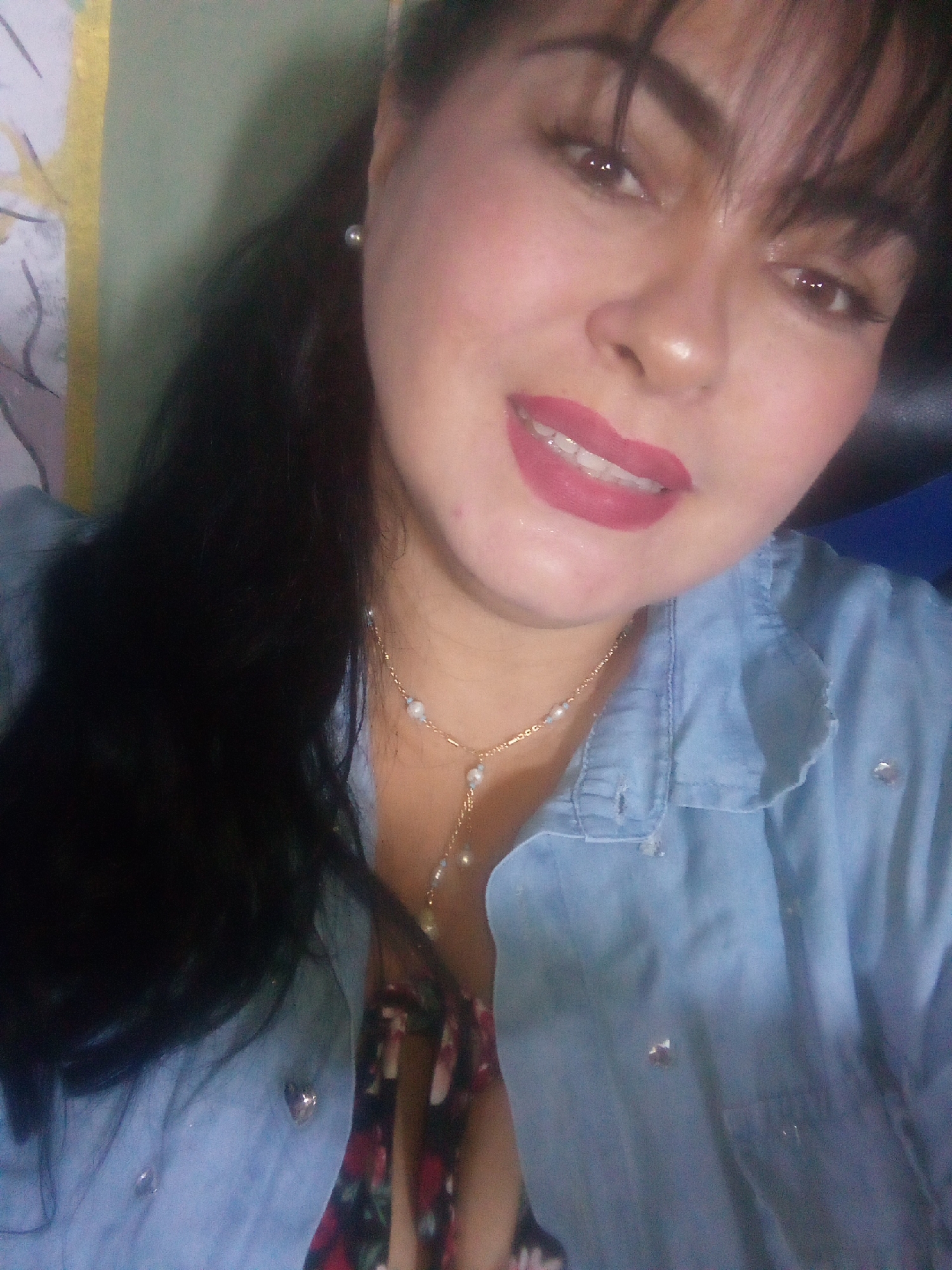 A woman with fair skin, dark hair, and a pearl earring is smiling at the camera. She is wearing a light blue denim shirt over a floral top and a delicate gold necklace with pearl accents. The location in the city of Juan Griego, Venezuela, is not discernible from the image.