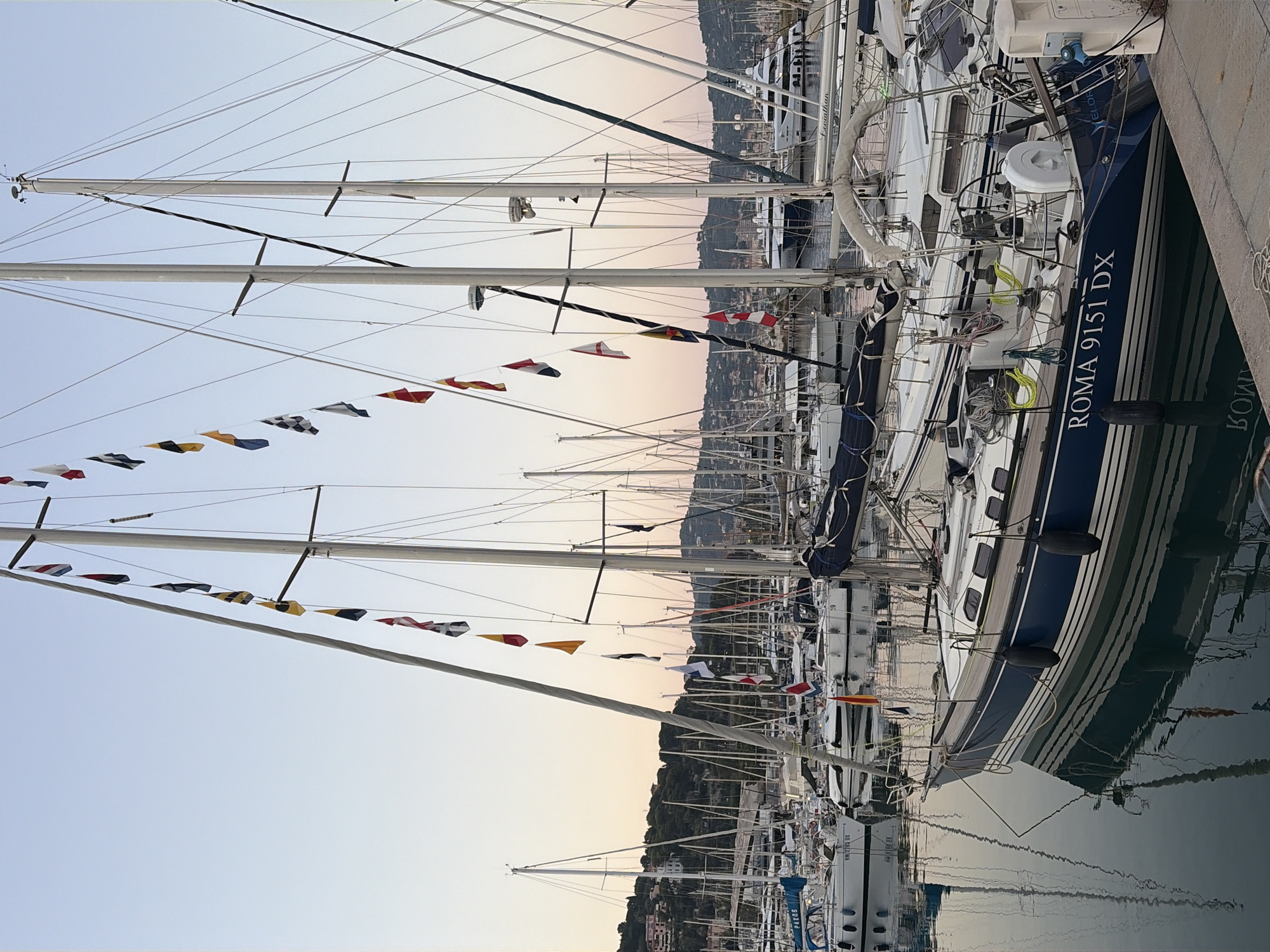 The image depicts a marina filled with sailboats at dusk. The sky displays a soft gradient of pale orange and pink, indicating sunset, with a clear and calm atmosphere. Numerous sailboats are moored alongside each other, their masts creating a dense forest of vertical lines against the sky. Many of the boats are decorated with strings of colorful signal flags, adding a festive element to the scene. One prominent sailboat in the foreground has the text "ROMA 9151 DX" visible on its hull. The boats are reflected in the still water of the marina, creating a mirrored effect that doubles the visual impact of the masts and flags. In the background, a town with buildings is nestled on a hillside overlooking the water, suggesting the location is Imperia, Italy. The overall mood is peaceful and serene, capturing the quiet beauty of a marina as day transitions to night.