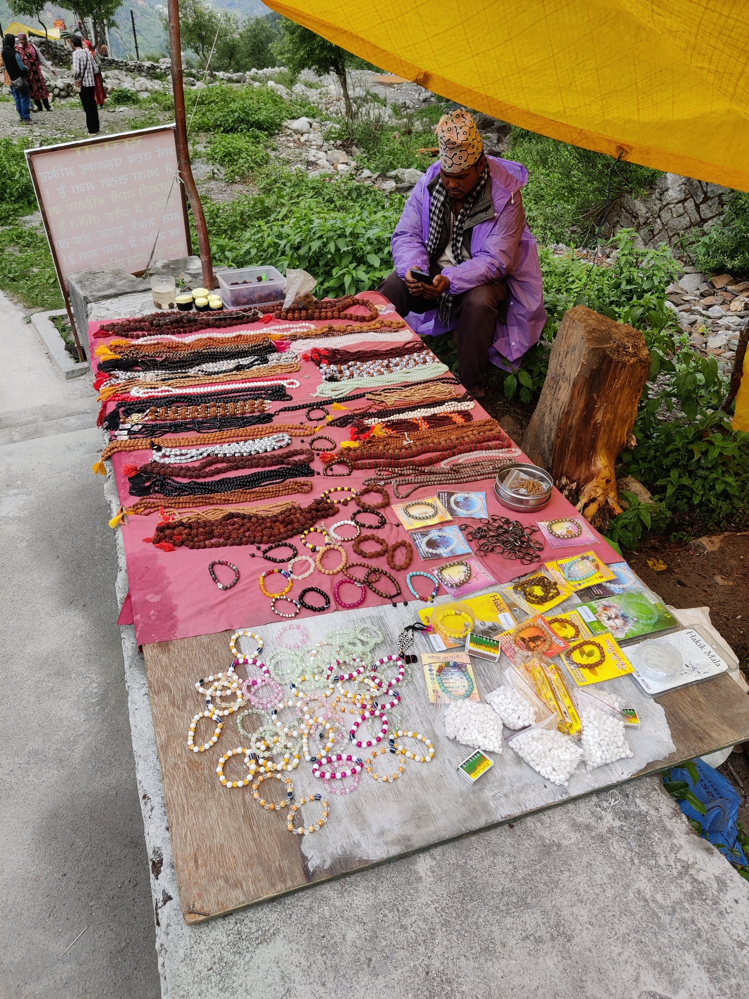 In Majhach, India, a street vendor sits under a yellow tarp, attending to his merchandise. The vendor, wearing a purple raincoat and a patterned cap, is engrossed in his mobile phone. His stall, draped with a pink cloth, displays an array of religious and decorative items. Primarily, the stall features numerous strings of beads, necklaces, and bracelets, made from various materials like wood, stone, and seeds, in a spectrum of colors and sizes. Some items are presented in small plastic bags, while others are loose. In the background, lush green foliage and rocky terrain suggest a natural, possibly mountainous, environment. Two individuals can be seen walking away from the stall in the distance, their figures partially obscured by the surroundings. A sign with writing in an Indian script is also visible. The overall scene captures a moment of commerce and daily life in a vibrant, outdoor setting.