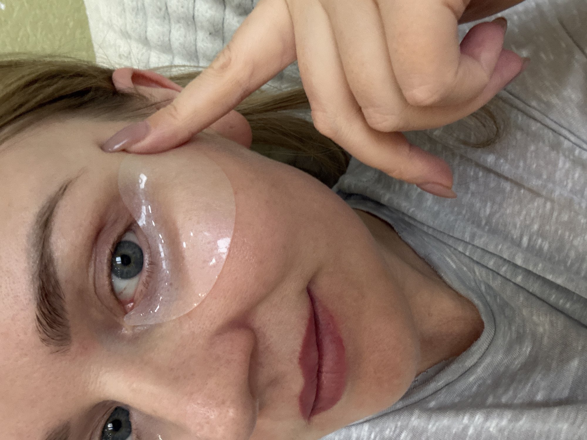 A woman is depicted indoors, engaged in a self-care routine, possibly in Chaikovsky, Russia. She is seen from a slightly elevated angle, lying down with her head resting on a light grey, textured fabric, likely bedding or clothing.
Clear, gel-like under-eye patches are applied beneath her eyes, with one prominently visible under her left eye, extending towards her temple. Her left hand, featuring manicured nails painted in a taupe or light brown shade, has its index finger gently touching her forehead near her left eyebrow, suggesting a natural pose or slight adjustment.
The woman has light brown hair, and her expressive blue-grey eyes are open and looking towards the viewer. Her lips show a soft, natural reddish tint, possibly from lip makeup or a natural flush. Her expression appears calm and neutral. The lighting suggests an indoor setting, likely during the day or evening with artificial light. There is no visible text in the image.