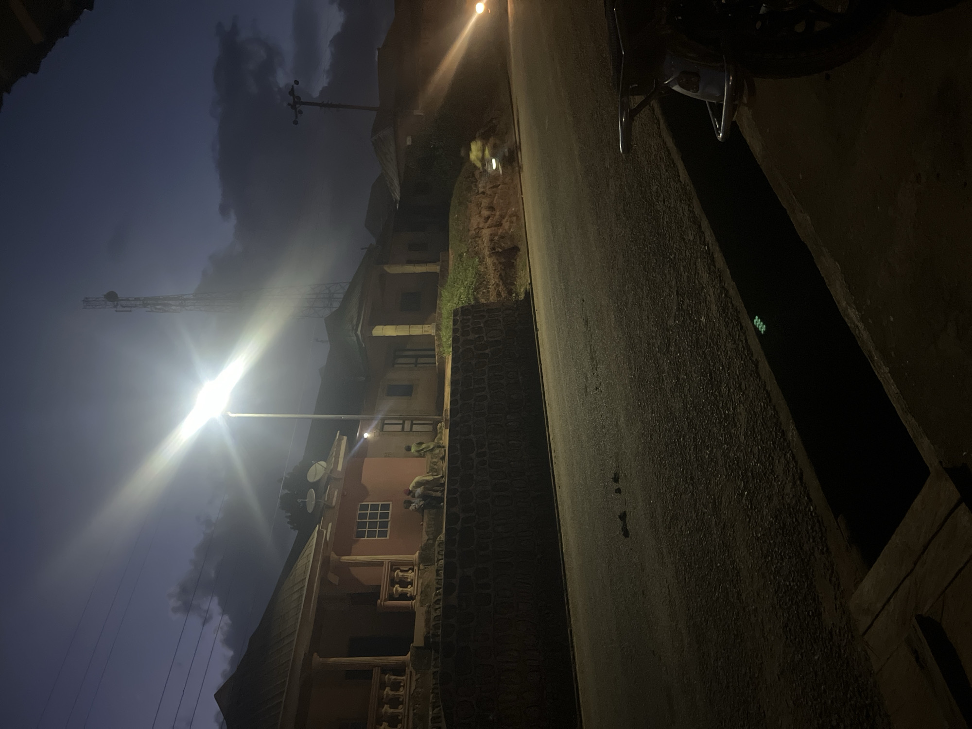 The image depicts a street scene in Gembu, Nigeria, at dusk or night. A bright street lamp illuminates the left side of the frame, casting strong light and lens flare. In the background, a tall telecommunications tower stands against a dark, cloudy sky. Several houses with pitched roofs are visible along the street, with some featuring satellite dishes. A low, dark stone wall lines the property adjacent to the road. A group of at least three individuals, dressed in traditional clothing, are gathered on top of this wall, near the houses. On the right side of the image, the headlights of a vehicle, possibly a motorcycle or car, are visible, creating a blurred streak of light on the road, indicating movement. A drainage ditch runs along the edge of the road, with a wooden plank partially covering it in the foreground. A portion of a motorcycle is visible in the lower right corner. The overall atmosphere is dim and moody, characteristic of a rural or semi-urban setting after sunset.