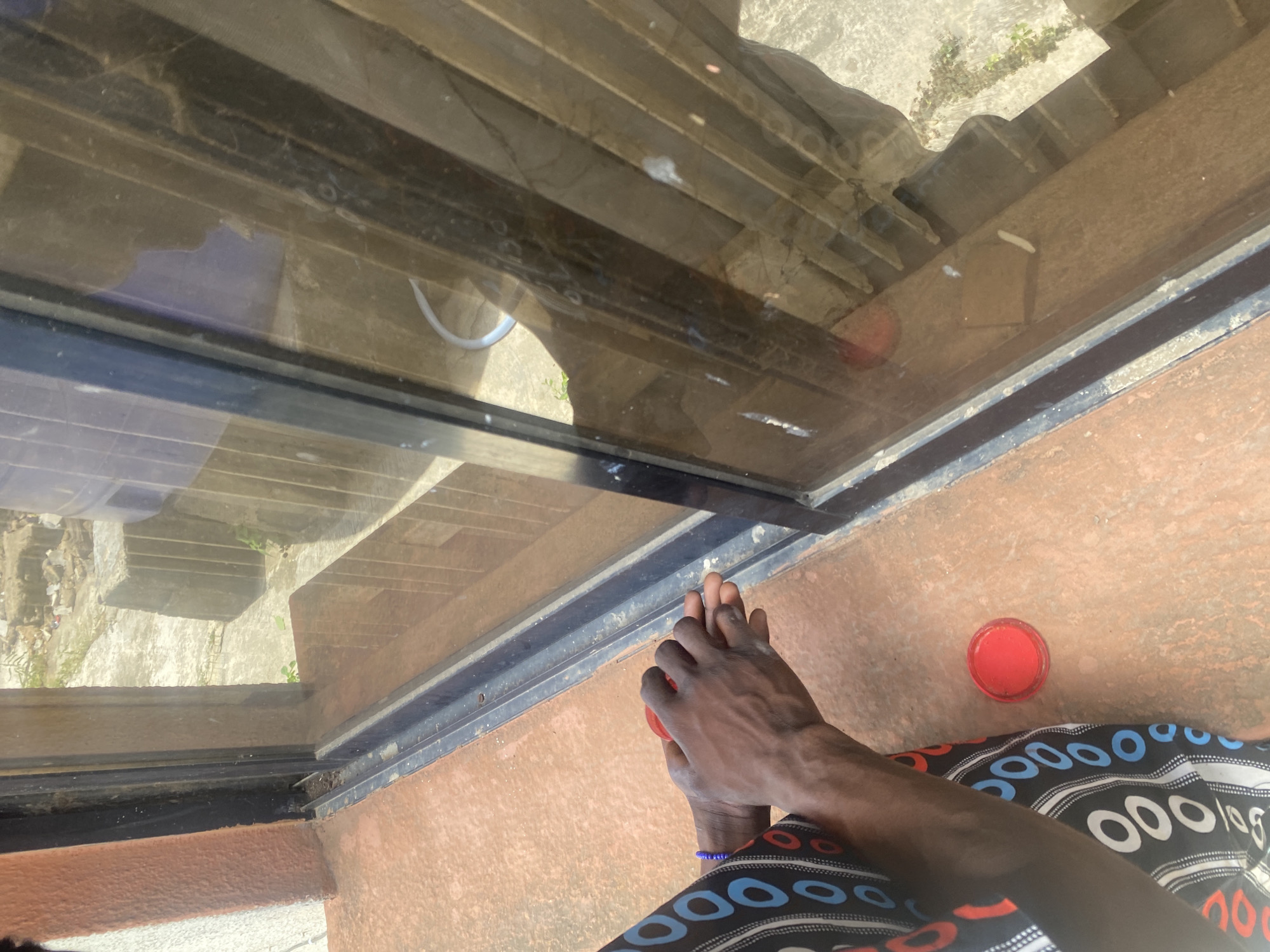 The image, taken from an elevated perspective, shows a person's hands holding a red object, possibly a small container. The person is wearing clothing with a black and white circular pattern. The background consists of a window reflecting an exterior scene with stairs and greenery. The surroundings appear to be a concrete or tiled surface. There is also another identical red object, and that the person is wearing a bracelet with blue beads. It is not possible to confirm the location in Lagos, Nigeria based solely on this image.
