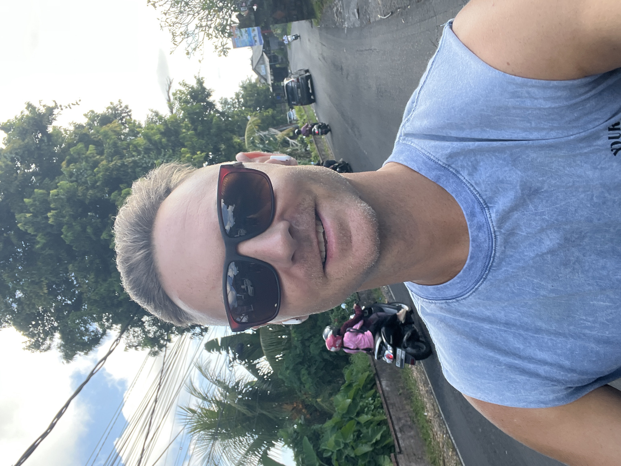 A man wearing sunglasses and a blue tank top is facing the camera, seemingly taking a selfie. He has short blonde hair and earbuds in his ears. Behind him is a street in Sukawati, Indonesia. Motorbikes are visible on the road, as well as a black car further away. Green trees and foliage are present, as well as power lines. A building with a banner is also visible in the background. The sky is partly cloudy.