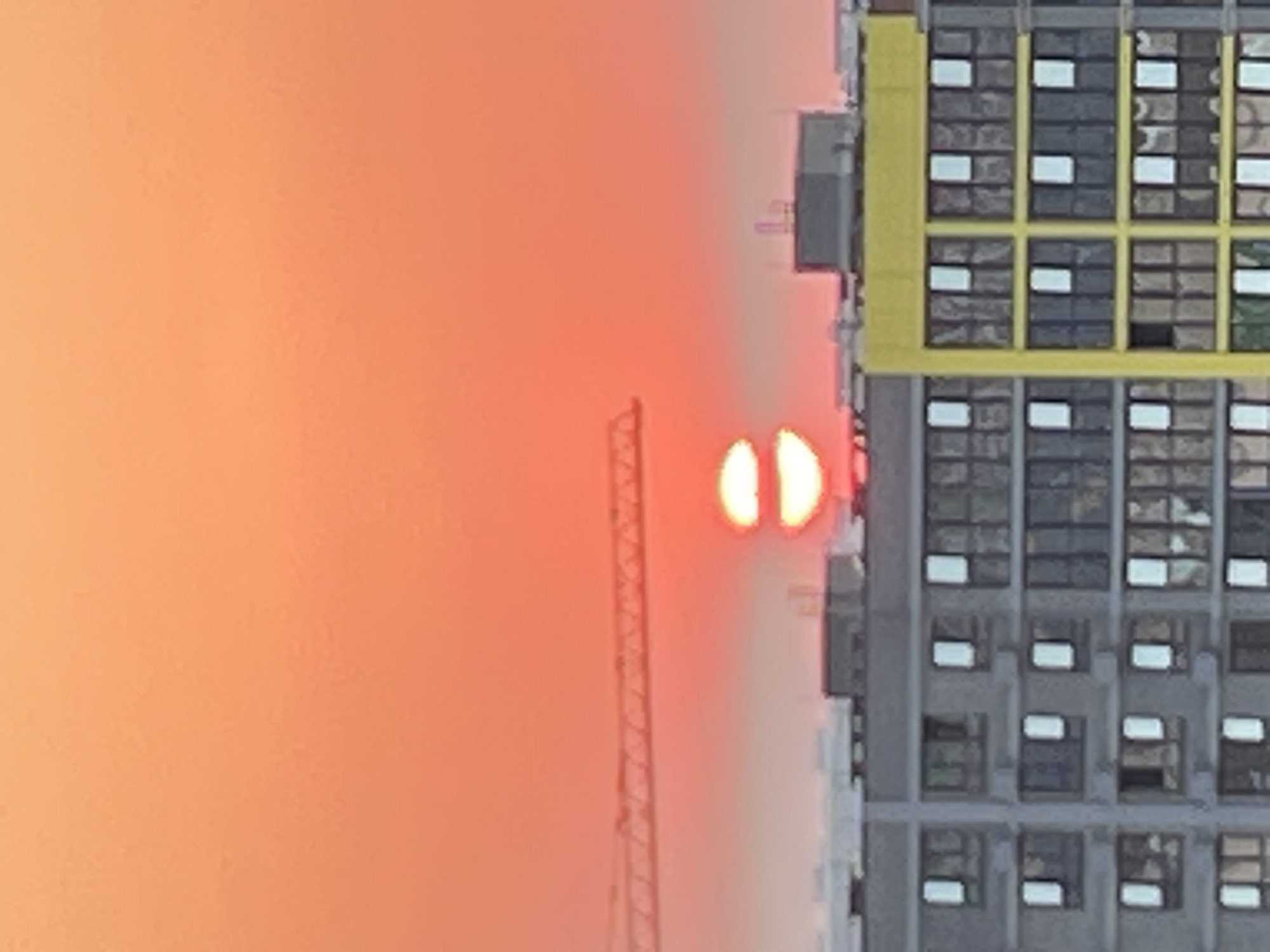 The image depicts a sunset over a cityscape, likely in Krasnodar, Russia, given the context. The sky is a vibrant gradient of orange and red, with the sun appearing as two distinct, bright discs stacked vertically, suggesting atmospheric distortion. In the midground, a construction crane's lattice structure stretches horizontally across the frame. Below the crane and sun, multiple multi-story buildings are visible. One building is predominantly gray with numerous windows, some of which are illuminated. Another adjacent building features a striking yellow facade with a grid of dark windows. The overall impression is that of an urban environment during dusk, with ongoing construction activity and residential or commercial structures. No people are discernible in the image.