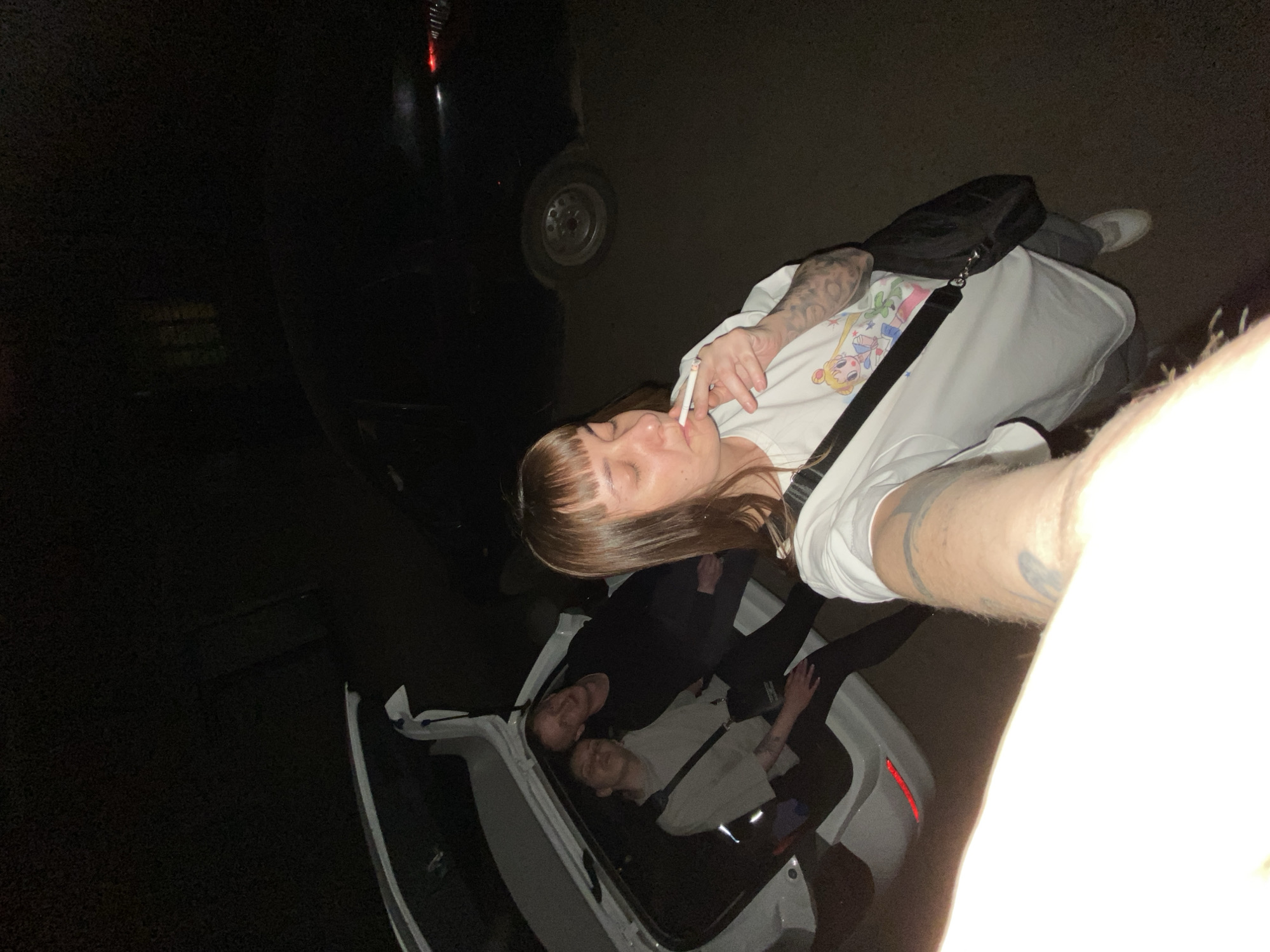 The image depicts an outdoor scene at night. A woman with brown hair and bangs is holding a cigarette in her mouth, wearing a white graphic t-shirt, gray pants, white shoes, and a black crossbody bag. She is angled toward the camera, possibly taking a selfie. The lower part of the frame shows the open trunk of a white car. Inside the car trunk, there are two other people, possibly women, lying down. One is dressed in black and the other in a lighter-colored top. A dark-colored car is also partially visible in the background. The location is potentially Votkinsk, Russia, based on the user's prompt.