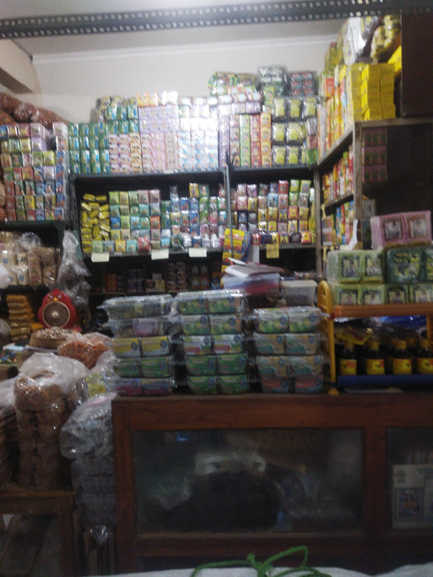 The image depicts a cluttered storefront in Sukoharjo, Indonesia, likely a small shop selling a variety of goods. Shelves are packed high with colorful boxes and packaged items, including what appear to be snacks or small food products. Some shelves are organized, while others are precariously stacked.

In the foreground, a wooden display cabinet with glass panels showcases more products. To the left, a red electric fan is visible, next to a round cake or pastry. Bags of food items, possibly crackers or similar snacks, are also stacked on a wooden surface. A green rope is visible in the bottom right corner. The lighting appears to be artificial and somewhat dim, contributing to the overall impression of a busy, perhaps slightly disorganized, retail environment. The scene is likely indoors, given the presence of shelving units and display cabinets.