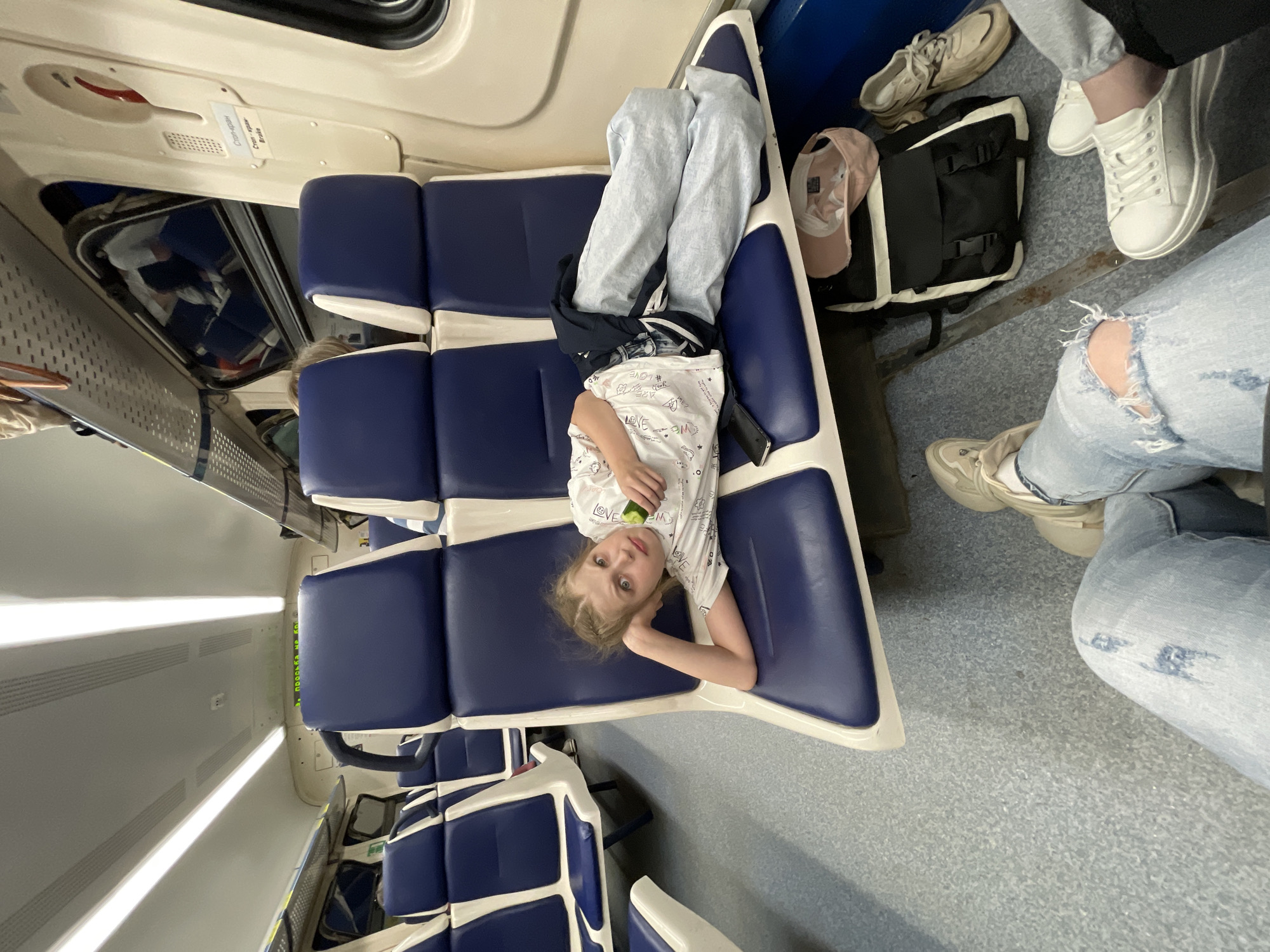 A young girl lies on a train seat in what appears to be a carriage. She is wearing a white shirt with patterns, light gray sweatpants, and appears to be holding a piece of green fruit or vegetable. Her head is resting on her hand.
Several other people's legs are visible near the edges of the frame; one person is wearing ripped light blue jeans and white sneakers. There's also a backpack and hat on the floor. The seats are blue with white trim, and the interior of the train is predominantly white and gray. The carriage has windows and lighting on the ceiling.
Based on the presence of signs in Russian in the cropped images, it is possible that this photograph was taken on a train traveling in or near the city of Agryz, Russia.