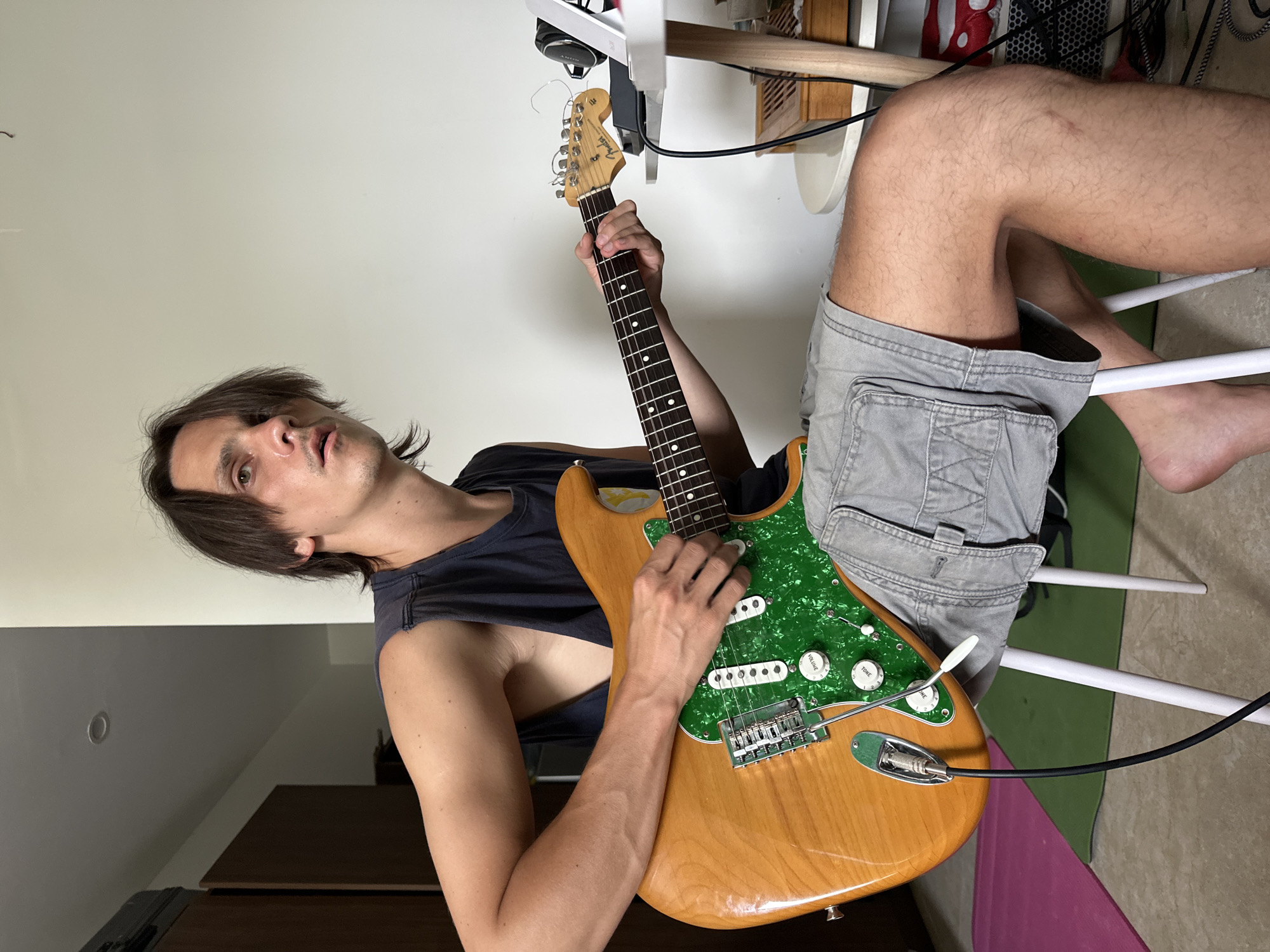 The image depicts a young adult male sitting on a white chair and playing a light-brown electric guitar with a green pickguard. He is wearing a dark sleeveless shirt and grey shorts. The guitar is plugged into a black cable. The background shows a white wall, a wooden wardrobe, and a white table with headphones and a tablet. The floor has a pink yoga mat and a green mat. The scene appears to be indoors, possibly a residential setting in Da Nang, Vietnam, given the available information about the location. The man is looking towards the left of the frame with an intent expression.