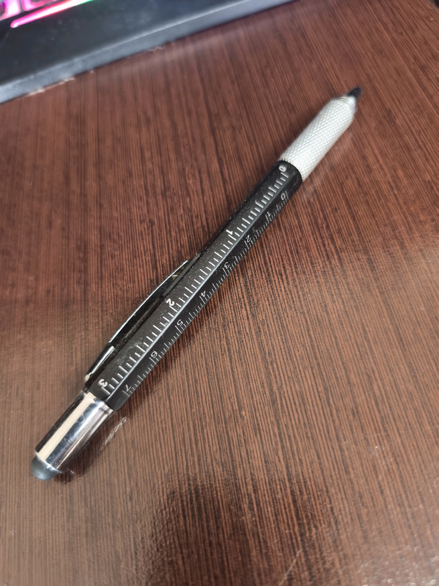A black multi-tool pen with a silver stylus tip rests diagonally on a brown wooden surface. The pen is inscribed with ruler markings in white. The background shows a portion of a black object with colorful lights. The location is Karachayevsk, Russia.