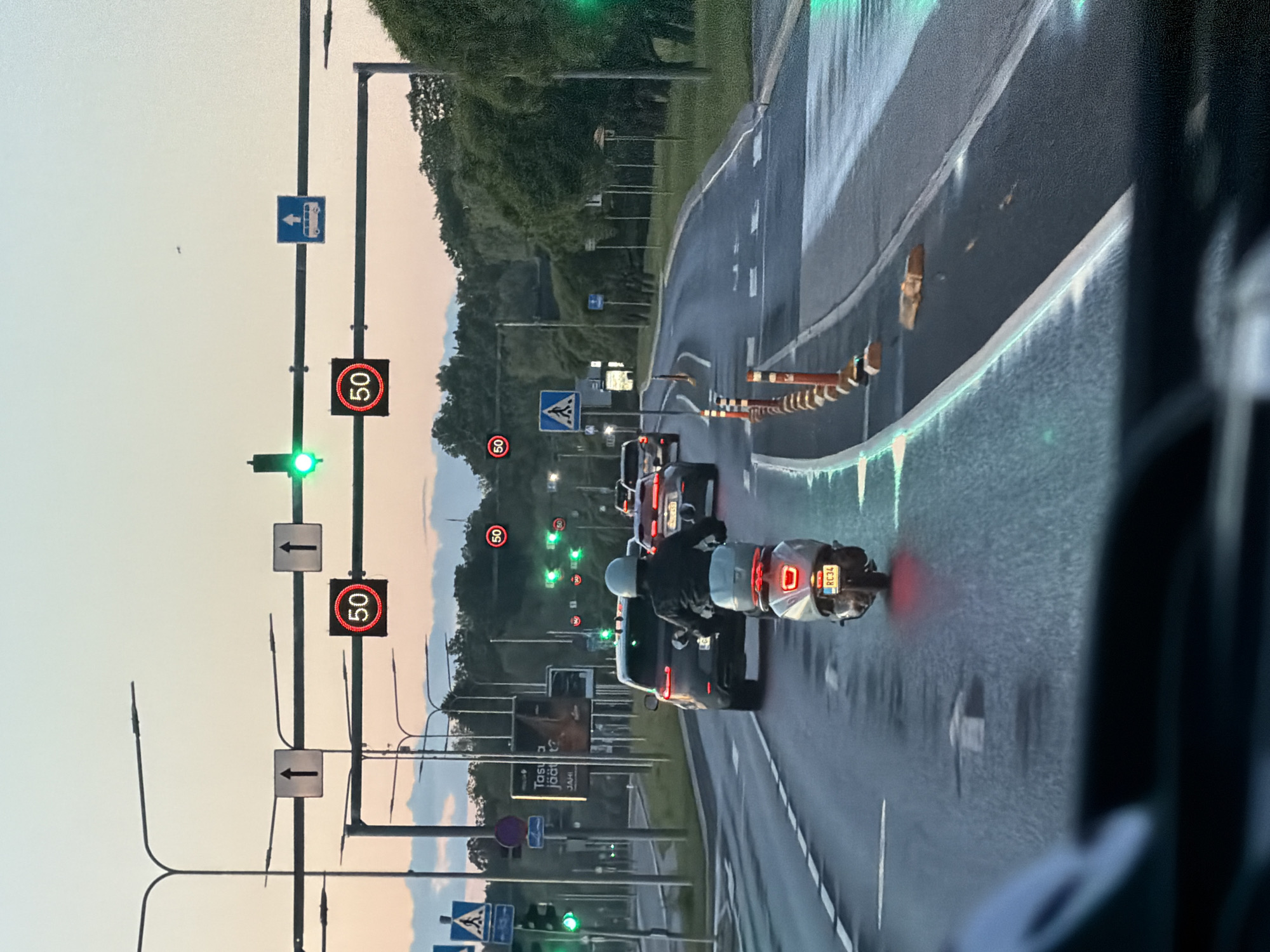 The image shows a street scene in Tallinn, Estonia, with traffic moving along a multi-lane road. In the foreground, a person is riding a scooter. Behind the scooter, several cars are visible, including a dark SUV and a silver sedan. Traffic lights are present, with some showing green signals. Speed limit signs indicating "50" are also visible. The environment appears to be a city street with trees lining the sides of the road. The sky is a pale orange and blue, suggesting it might be dusk or dawn. Road markings are clear, and there are some temporary traffic cones on the right side of the road. The overall scene depicts ordinary urban traffic flow.