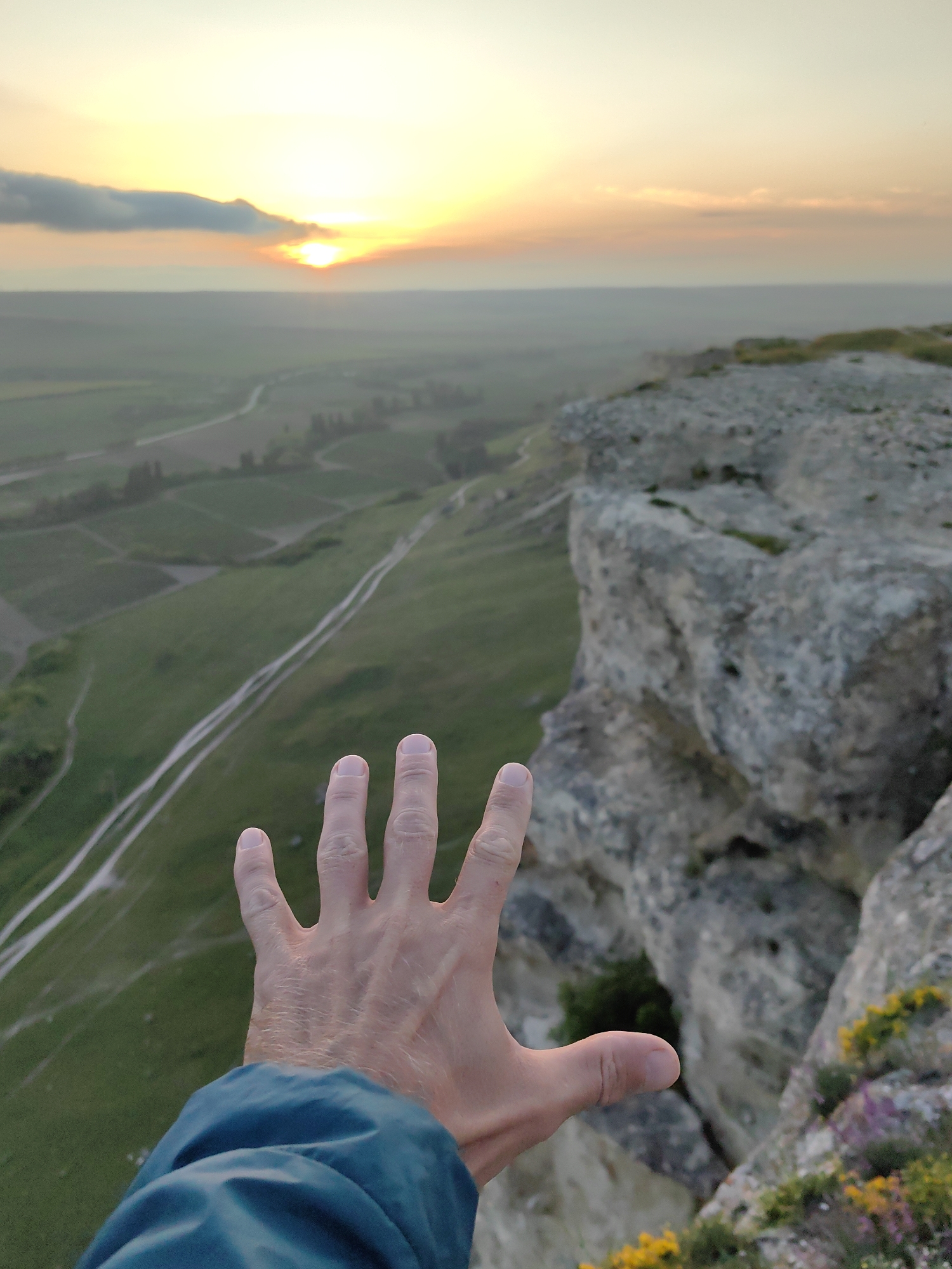 The image shows a hand outstretched towards a sunset view from a cliff edge. The foreground is dominated by a person's hand, with the wrist and a portion of a blue jacket sleeve visible. The hand is open, with fingers spread.
The background reveals a vista of rolling green fields and valleys, dissected by white trails or roads. The land appears to be agricultural, with rows of crops or vineyards. In the distance, a sunset is partially obscured by a cloud, casting an orange glow across the sky.
The immediate foreground to the right of the hand includes the rocky edge of a cliff with some sparse vegetation. The location of the scene is Unknown, Unknown.