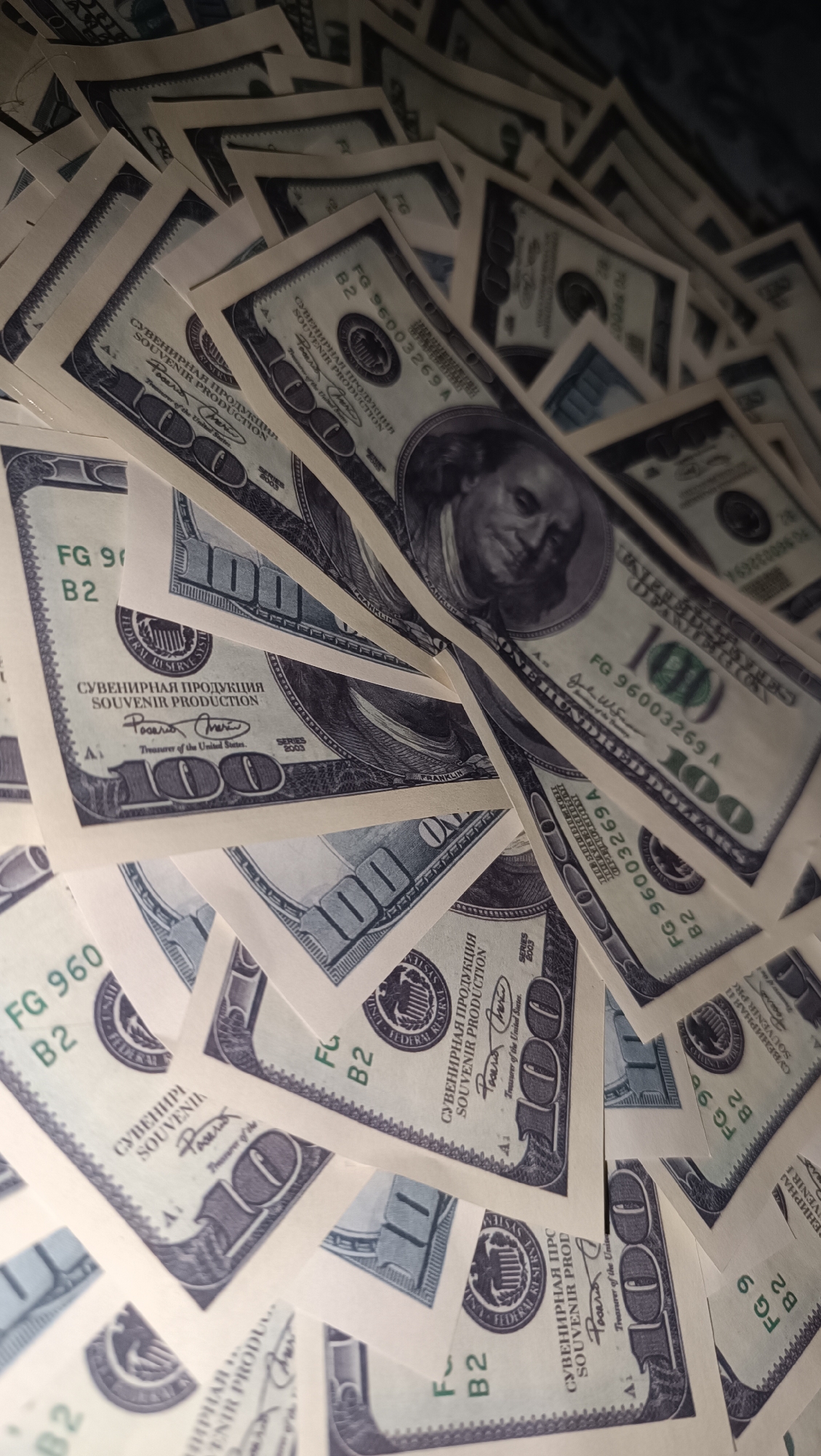 The image displays a close-up, overhead view of a pile of US one hundred dollar bills, predominantly featuring Benjamin Franklin's portrait and the number "100." The bills are arranged in a fan-like manner, overlapping each other, with some appearing crisp and new, while others show signs of wear. The lighting casts subtle shadows, highlighting the texture and details of the currency. The image is cropped tightly, focusing solely on the money, and provides no contextual information about its location in Chaikovsky, Russia, or any other city. There are no people, actions, or interactions depicted in the scene. The environment is not discernible beyond the surface on which the money is placed.