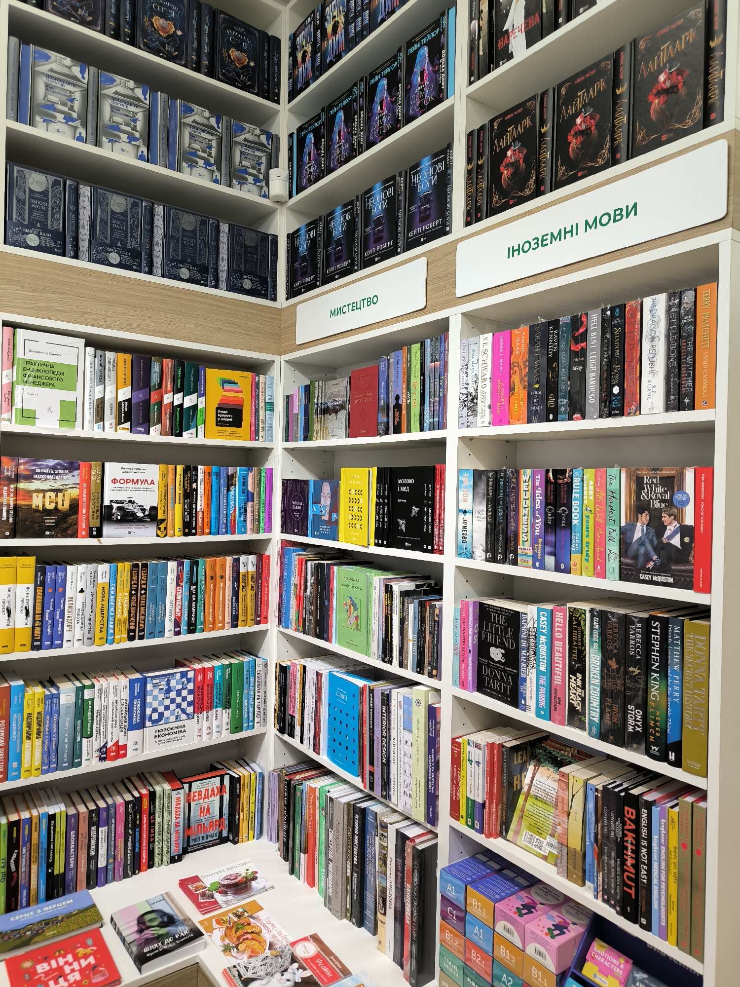 The image shows the interior of a bookstore, potentially located in Vinnytsia, Ukraine, given the use of Ukrainian language on book covers and section labels. The shelves are tightly packed with books, categorized under headings like "Art," "Foreign Languages," and others primarily in Ukrainian. The books are displayed spine-out. There are a variety of book topics including business, finance, literature, fiction, self-help, and language learning. The lighting is bright, and the shelves are white with light wood accents. Some books are also laid out on a table at the front. The store appears well-stocked and organized. No people are visible in the image.