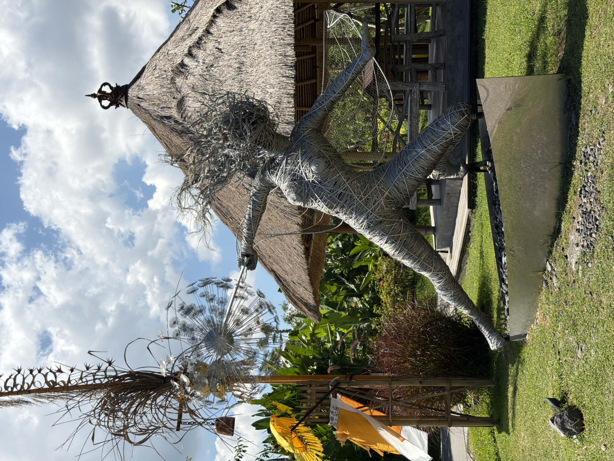 A wire sculpture of a human figure in a dynamic pose stands on a grassy lawn. The sculpture is made of twisted gray wire, creating a textured effect. The figure appears to be mid-movement, with its arms extended and its head turned. In the background, there is a thatched-roof structure, possibly a gazebo, with wooden furniture visible inside. Tall green plants and trees surround the area, and a bright blue sky with fluffy white clouds is overhead. Yellow and white fabric flags are attached to a wooden pole to the left of the sculpture. The scene is outdoors, suggesting a park or garden setting.