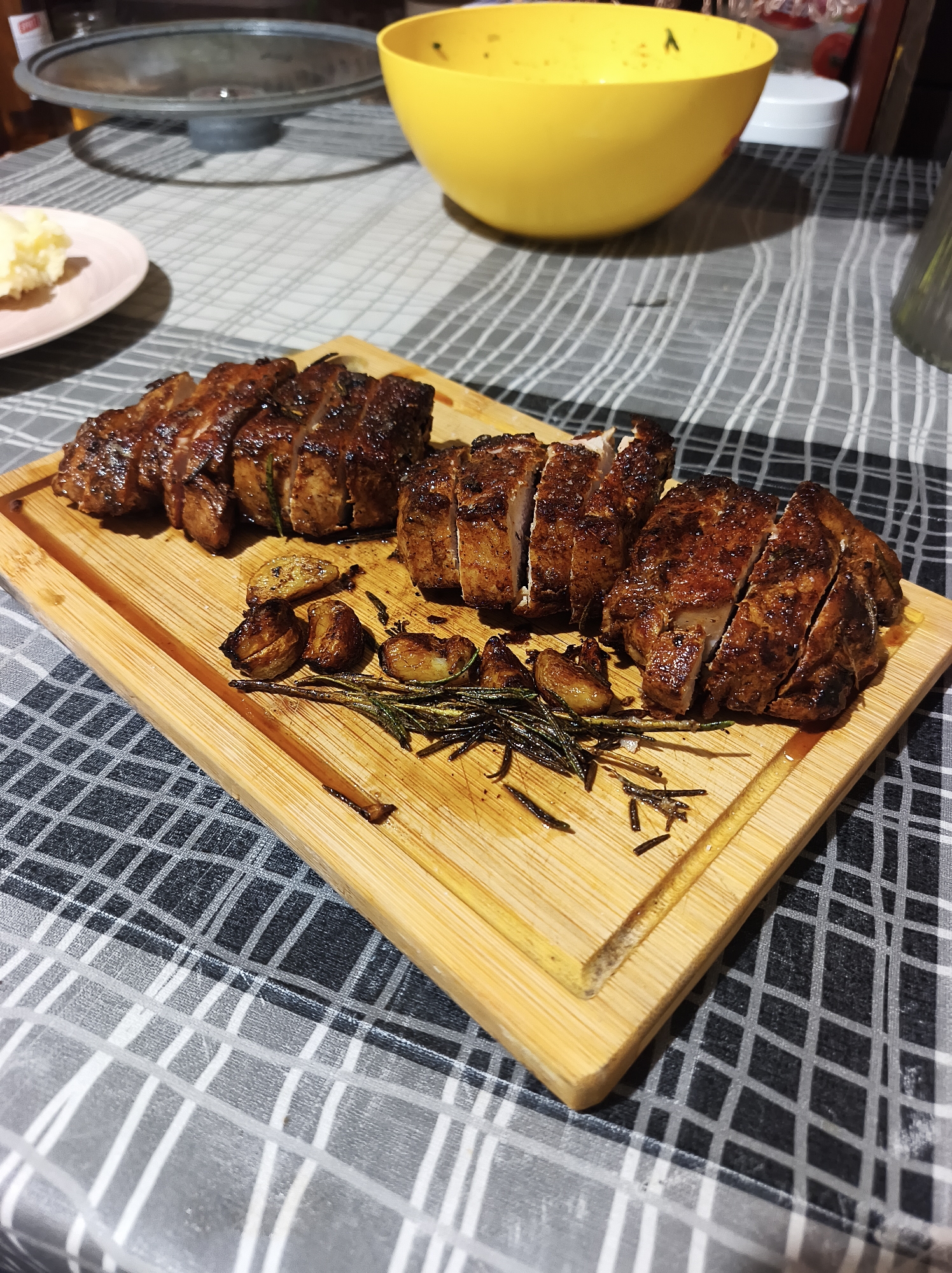 The image shows a wooden cutting board with sliced, roasted meat, garlic cloves, and rosemary sprigs. The cutting board is placed on a checkered tablecloth. In the background, there is a plate with mashed potatoes, a pot lid, and a yellow bowl. The location in the city of Ternopil, Ukraine is not evident. The image focuses on the food arrangement on the cutting board. There are no people visible.