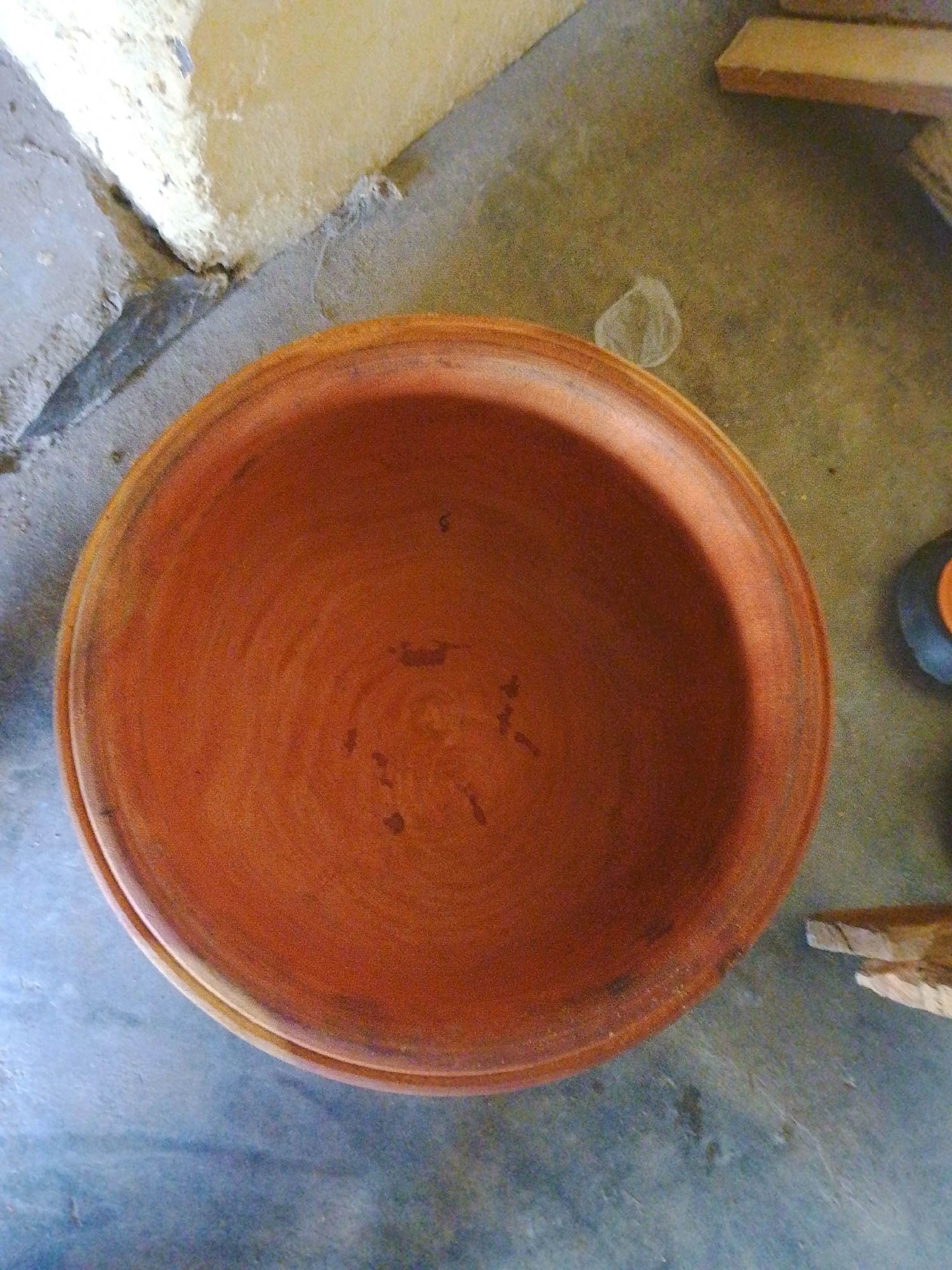 An overhead, medium shot captures a terracotta pot resting on a rough, concrete floor in an outdoor setting. The pot is round with a wide opening and appears to be used for cooking or storage. Its earthy orange hue is rich, with subtle variations in tone and texture visible on its surface. To the left of the pot, a portion of a light-colored, textured wall is visible, suggesting it might be an exterior or a section of a building. The floor beneath the pot is a mottled gray, interspersed with dirt and debris. To the right, the edge of a wooden plank can be seen, along with a glimpse of another object, possibly another pot or container. The overall scene is simple and utilitarian, evoking a sense of everyday life in a non-urban environment.