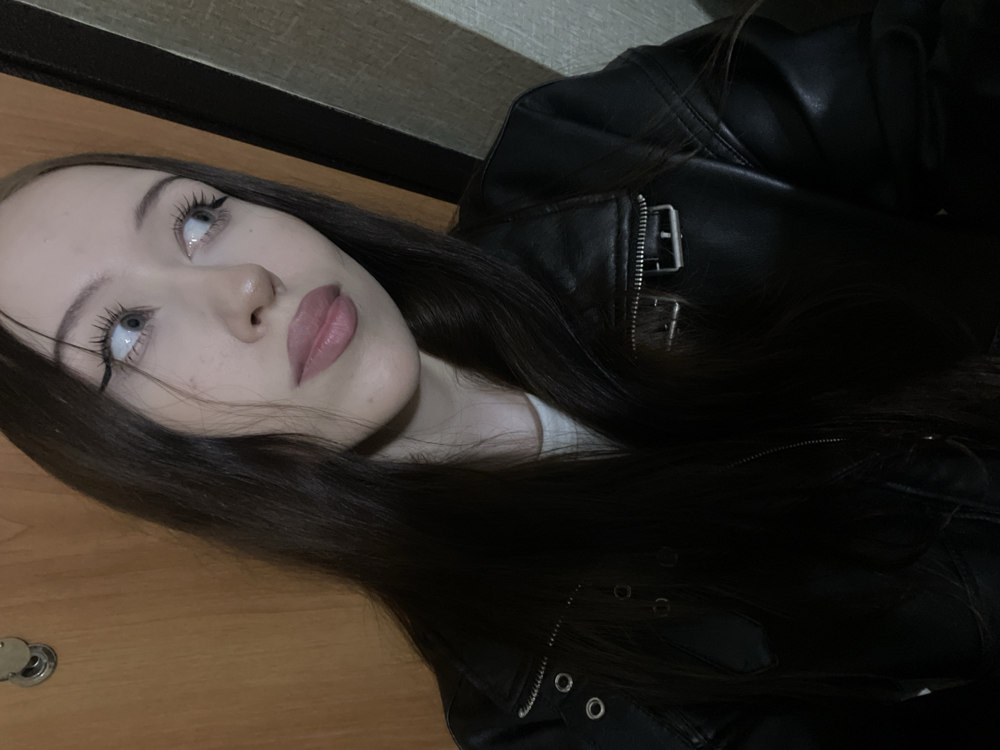 The image is a medium shot of a young woman with long, dark hair, wearing a black leather jacket. She is looking upwards and to the left, with a contemplative expression. Her eyes are wide open, and her lashes are long and dark. She is wearing subtle makeup, including eyeliner and mascara, and her lips are a soft pink. The background is blurred, but appears to be an indoor setting with a wooden surface and a textured wall. A doorknob is visible on the lower left side of the frame. The lighting is dim, suggesting it might be evening or nighttime. The overall mood of the image is introspective and somewhat dramatic. There is no visible text in the image.