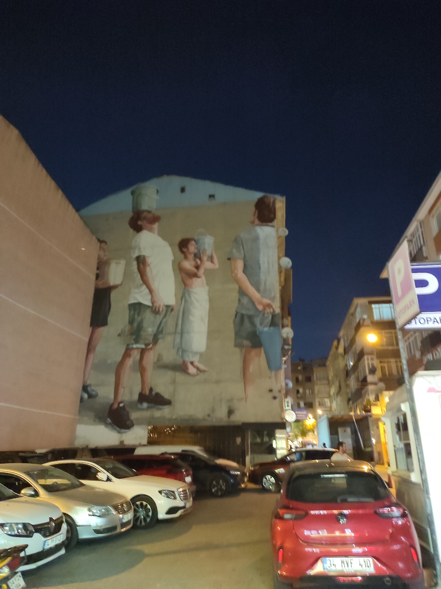 The image depicts a street scene at night in Kadıköy, Turkey, featuring a large mural on the side of a building. The mural shows three figures in what appears to be an artistic representation of carrying buckets. One figure on the left is partially obscured by a beige wall and is holding a white rectangular object. The central figure, a man, is wearing a white t-shirt, camouflage shorts, and dark sneakers, with a bucket balanced on his head. To the right, another man, shirtless and wearing a white towel around his waist and white pants, holds a bucket. In the foreground, several parked cars are visible. A red hatchback is in the immediate foreground, with a license plate reading "34 MVF 410". To its left, a silver sedan and a white sedan are parked. In the background, more cars and multi-story buildings are illuminated by streetlights. A "PARK" sign with a "P" symbol is mounted on a pole to the right of the frame. The sky is dark, indicating nighttime.